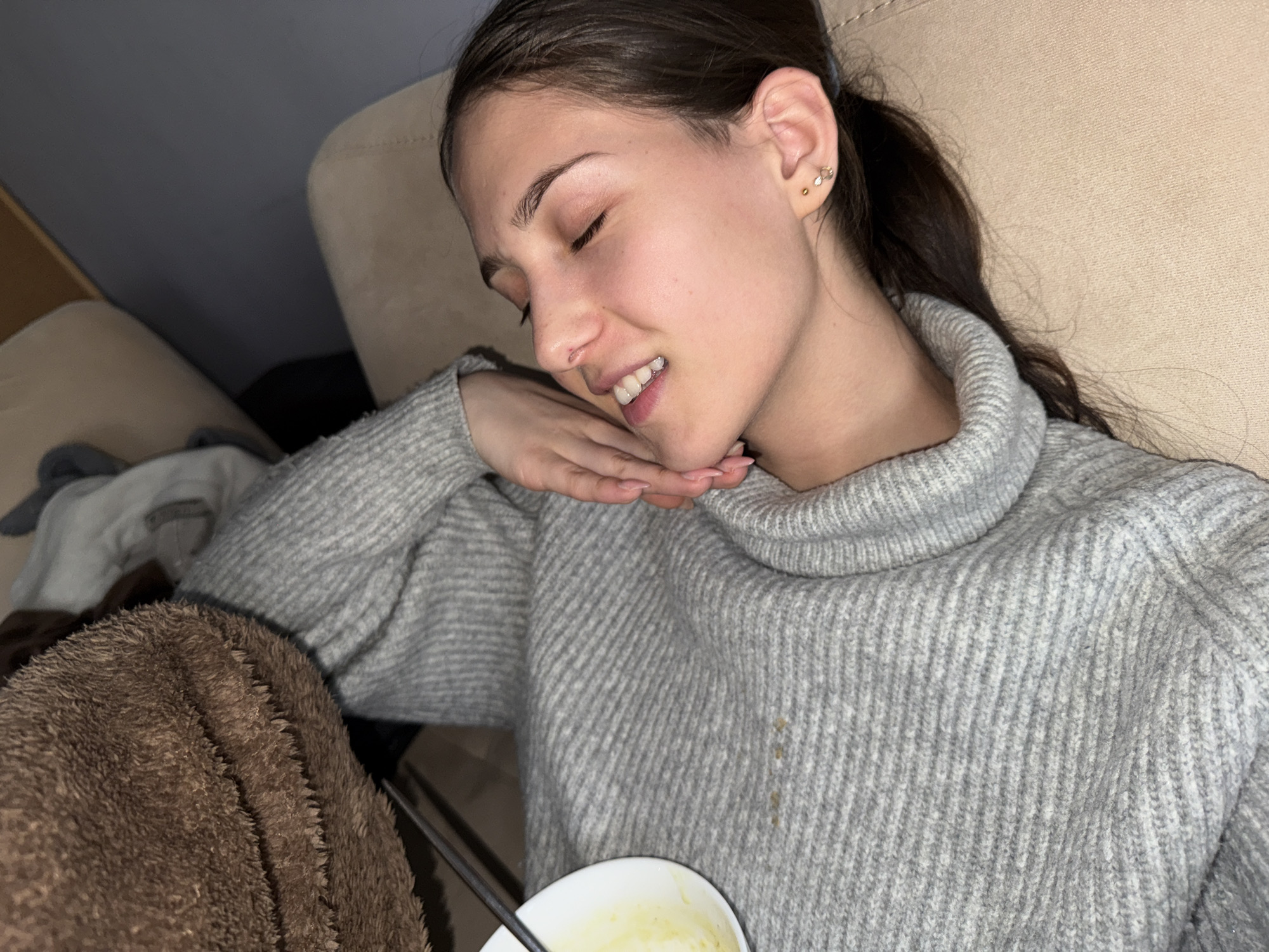 The image captures a young woman relaxing indoors, likely in Sofia, Bulgaria. She has dark hair pulled back in a ponytail and is wearing a light grey, ribbed turtleneck sweater. Her eyes are closed, and a subtle smile suggests a state of peace, contentment, or light sleep. Her head is gently resting on her right hand, which is visible with light pink nail polish.
In her left hand, partially visible at the bottom of the frame, she holds a white bowl containing a light-colored, possibly creamy food or drink, with a metal spoon inside. A fluffy brown blanket covers part of her lower body, contributing to the cozy atmosphere. She appears to be reclining on a light-colored sofa or couch. Two small stud earrings, one a cluster and one a gold bead, are visible on her right ear. There is a faint, light brown stain on the front of her sweater. The soft, warm lighting indicates an indoor setting, likely during the evening or night. The overall scene conveys a feeling of comfort and quiet relaxation. There is no visible text in the image.