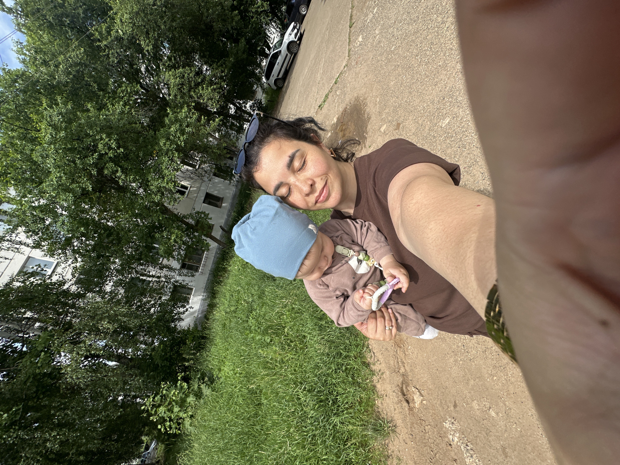 A woman in a brown t-shirt holds a baby wearing a blue hat and a brown onesie. The baby holds a pink toy in their hands. The woman has dark curly hair and sunglasses on her head. They are outdoors, with a grassy area to the left and a paved area with cars to the right. A building is visible in the background. The scene appears to be a residential area in a city.