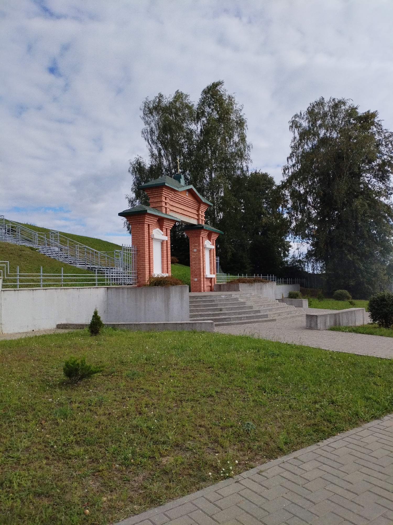 The image depicts an outdoor scene in Bialynichy, Belarus, featuring a brick archway with a green roof, likely an entrance to a memorial or religious site. In the background, a large hill is ascended by a wide staircase with metal railings. The archway itself is adorned with white, framed rectangular structures on either side. The surrounding area consists of manicured green lawns, a paved walkway, and several trees. The sky is partly cloudy with some blue visible. A small evergreen shrub is planted in front of the archway.