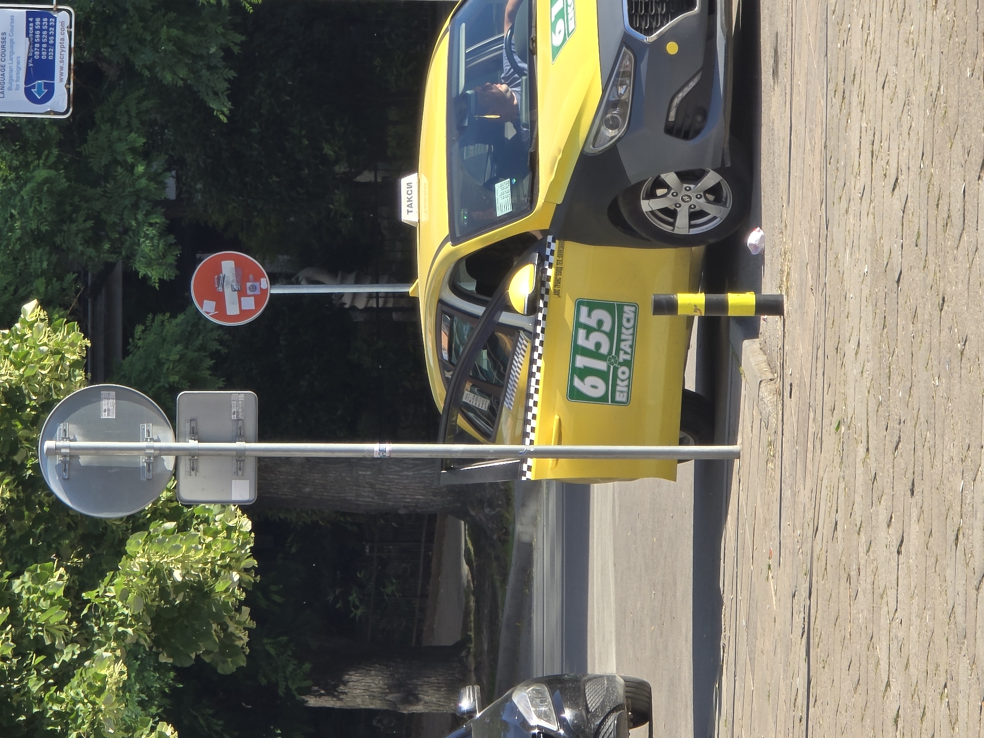 A bright yellow taxi cab with dark gray front quarter panels sits on a street in an unidentified city. The cab displays "6155 EKO TAKSI" in green lettering. A small sign reading "TAKCИ" is placed on top of the cab. Checkered trim runs along the door frame. Traffic signs are visible on a pole near the car. A sign for language courses, including website address, is posted nearby. Cobblestone pavement stretches across the foreground. The driver of the taxi is partially visible through the windshield. Another vehicle sits behind the taxi.