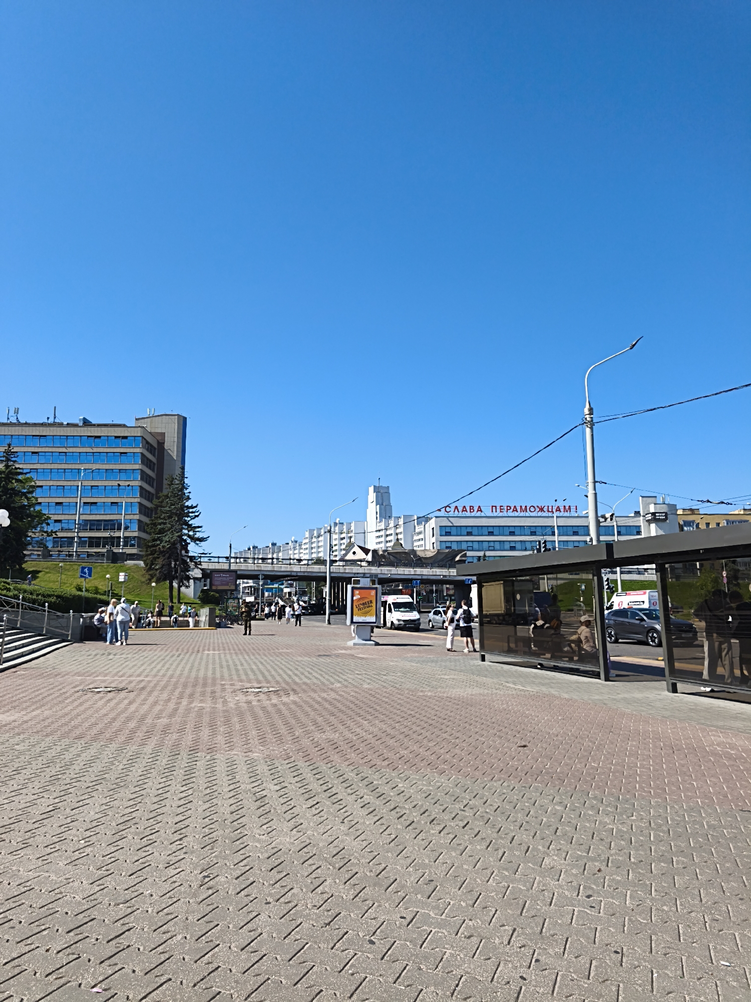 The image depicts a street scene in Minsk, Belarus, under a clear blue sky. In the foreground, a red and gray brick-patterned walkway stretches across the frame, leading towards the midground. To the left, a modern, multi-story blue-glass building stands prominently, bordered by a grassy slope and trees. A set of stairs descends from the left edge of the image towards a cluster of people standing near a subway entrance.
In the midground, a bus stop shelter with a glass enclosure is visible on the right. People are seated inside the shelter and standing outside. Across the street, there are cars and a white van. A prominent red sign with white Cyrillic text, reading "СЛАВА ПЕРАМОЖЦАМ!" (Glory to the Victors!), is displayed on a building in the background. Further back, more white buildings and an overpass are visible, suggesting an urban environment. The overall atmosphere is bright and sunny, with a moderate amount of pedestrian and vehicular activity.