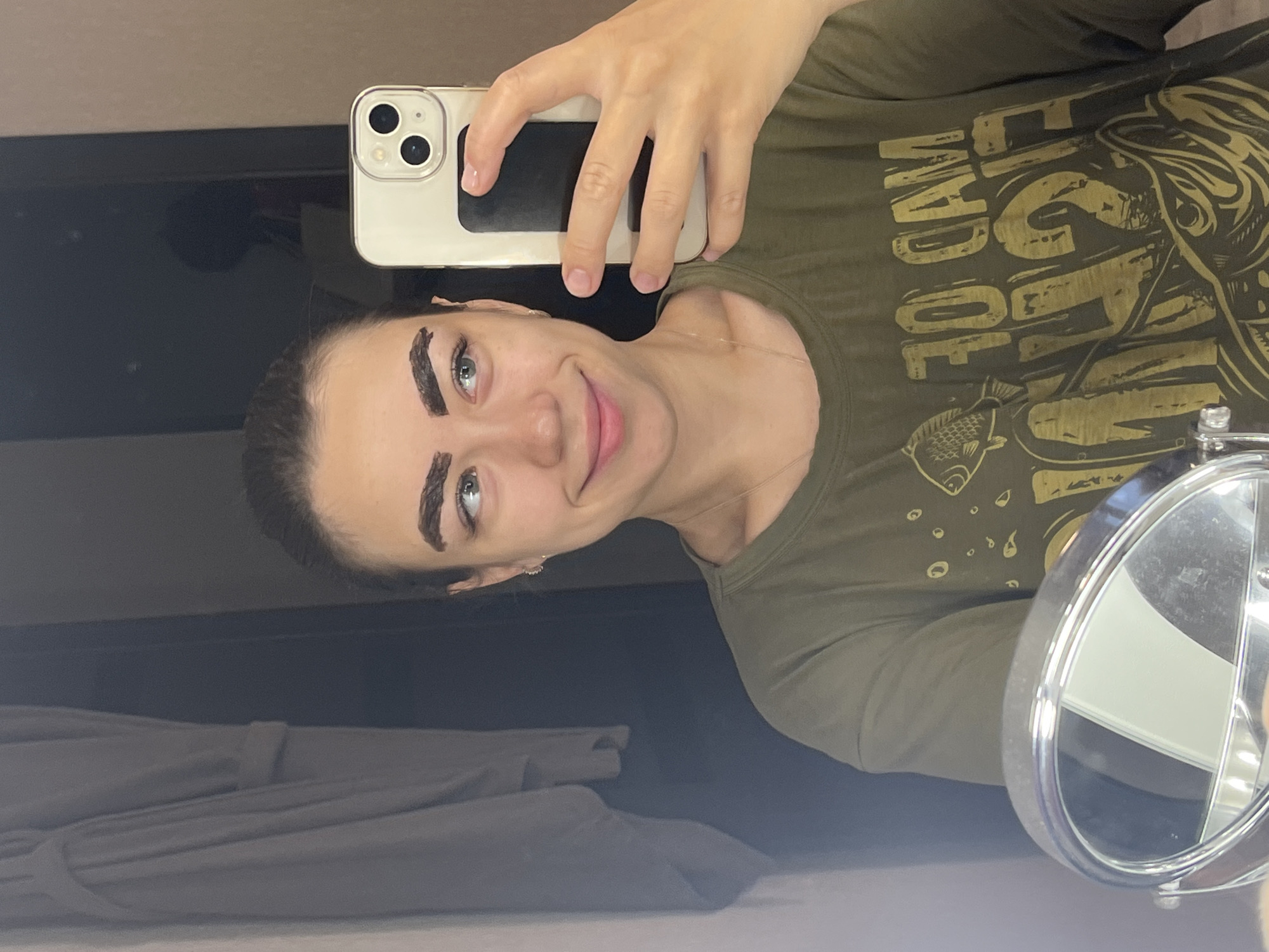 The image shows a young woman with dark, freshly dyed eyebrows, taking a selfie in a mirror. She is wearing a green t-shirt with a graphic of a fish and the words "MAD OF FISHING" printed on it. She is smiling and looking to her left. A makeup mirror is visible in the foreground. The background appears to be a bathroom, with a gray robe hanging on a door and a dark wall. The image does not provide enough context to determine the specific location in Chaikovsky, Russia, beyond it being an indoor setting likely in a private residence.