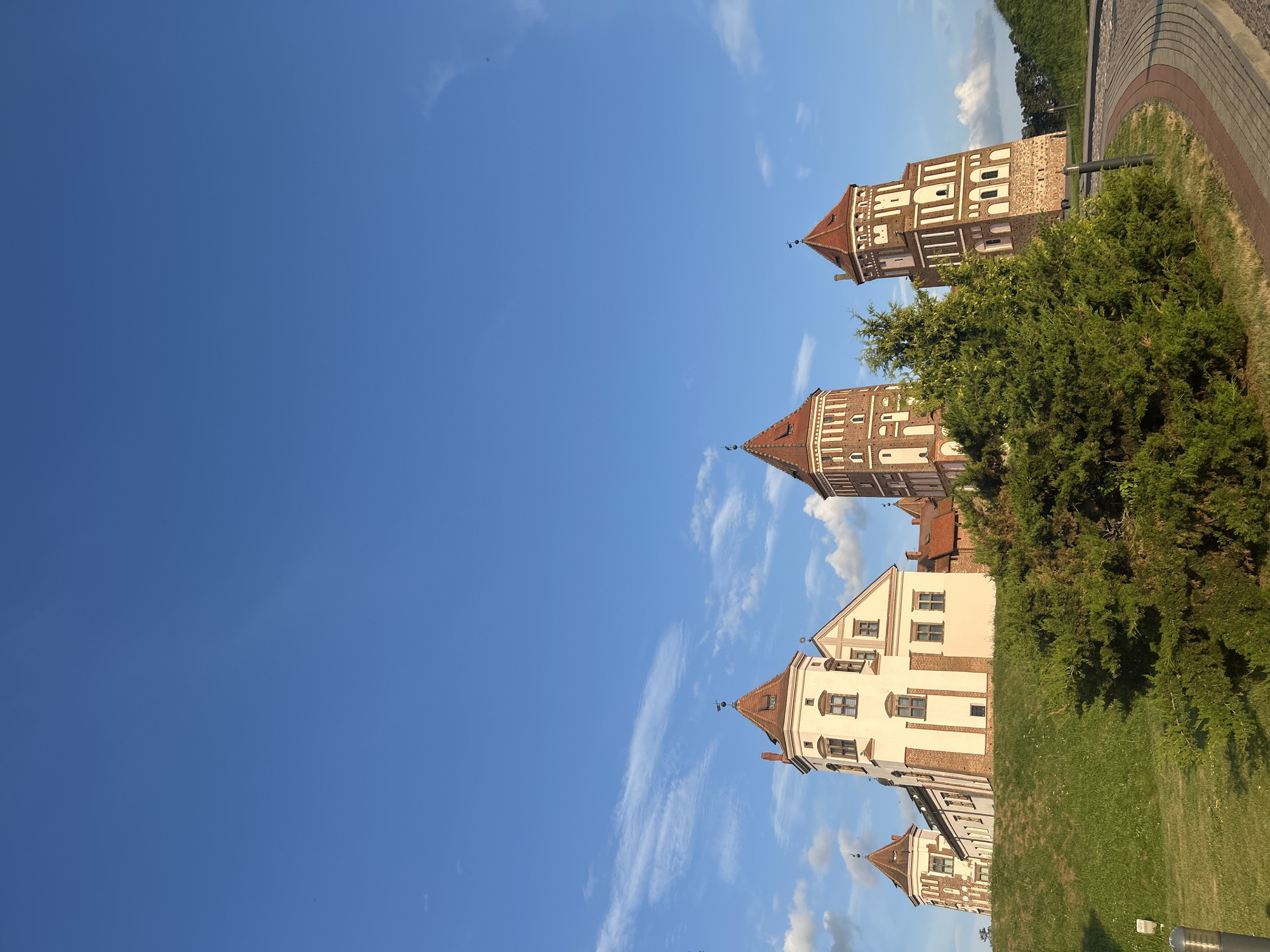 A daytime, eye-level, low-angle exterior shot captures a castle complex against a bright blue sky with scattered white clouds. The castle, located in Zhukhavichy rural council, Belarus, features multiple towers with red tiled roofs and light-colored walls. Some towers are constructed of red brick with white decorative elements, while others have white facades with brick accents. Green grass covers the sloping ground in front of the castle, with a mature evergreen shrub partially obscuring the lower portion of the scene. A paved pathway with red and grey brickwork curves into the foreground from the right, and a lamppost is visible on the left. The sun illuminates the castle, casting some shadows and highlighting the architectural details. There are no people or animals present.