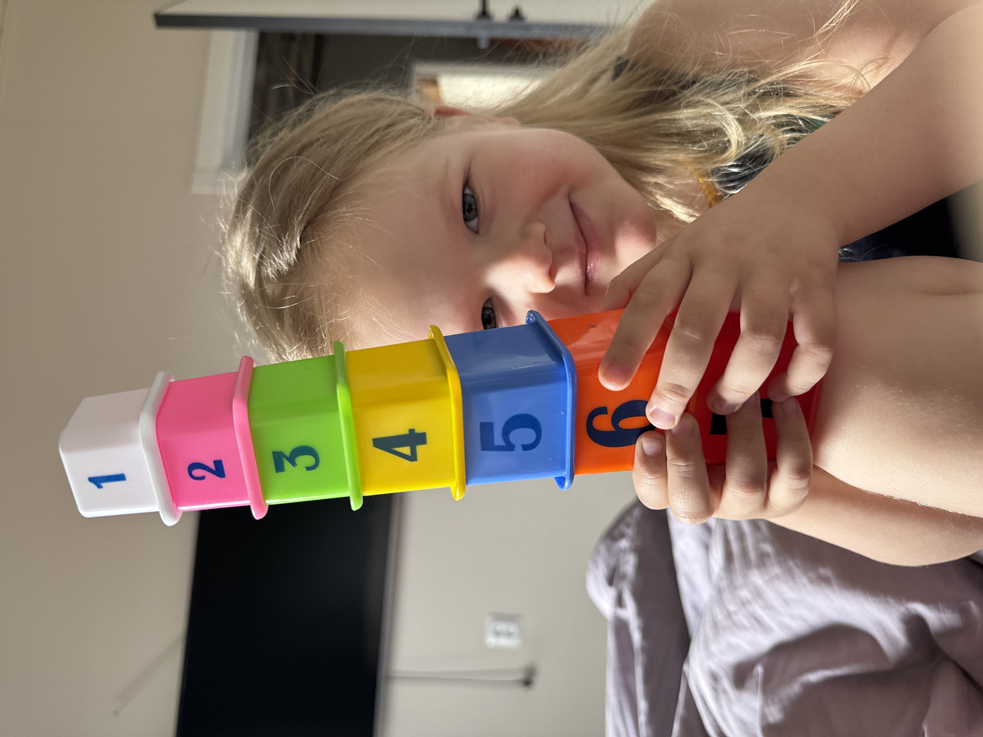 A young girl with blonde hair is holding a stack of colorful numbered cups. The cups are stacked in order from 1 to 6, with the colors being white, pink, green, yellow, blue, and orange. The girl is smiling and looking at the camera. The background is blurry, but it appears to be a room with a TV and light-colored walls. The location is not discernible to be in the city of Ферма, Russia.