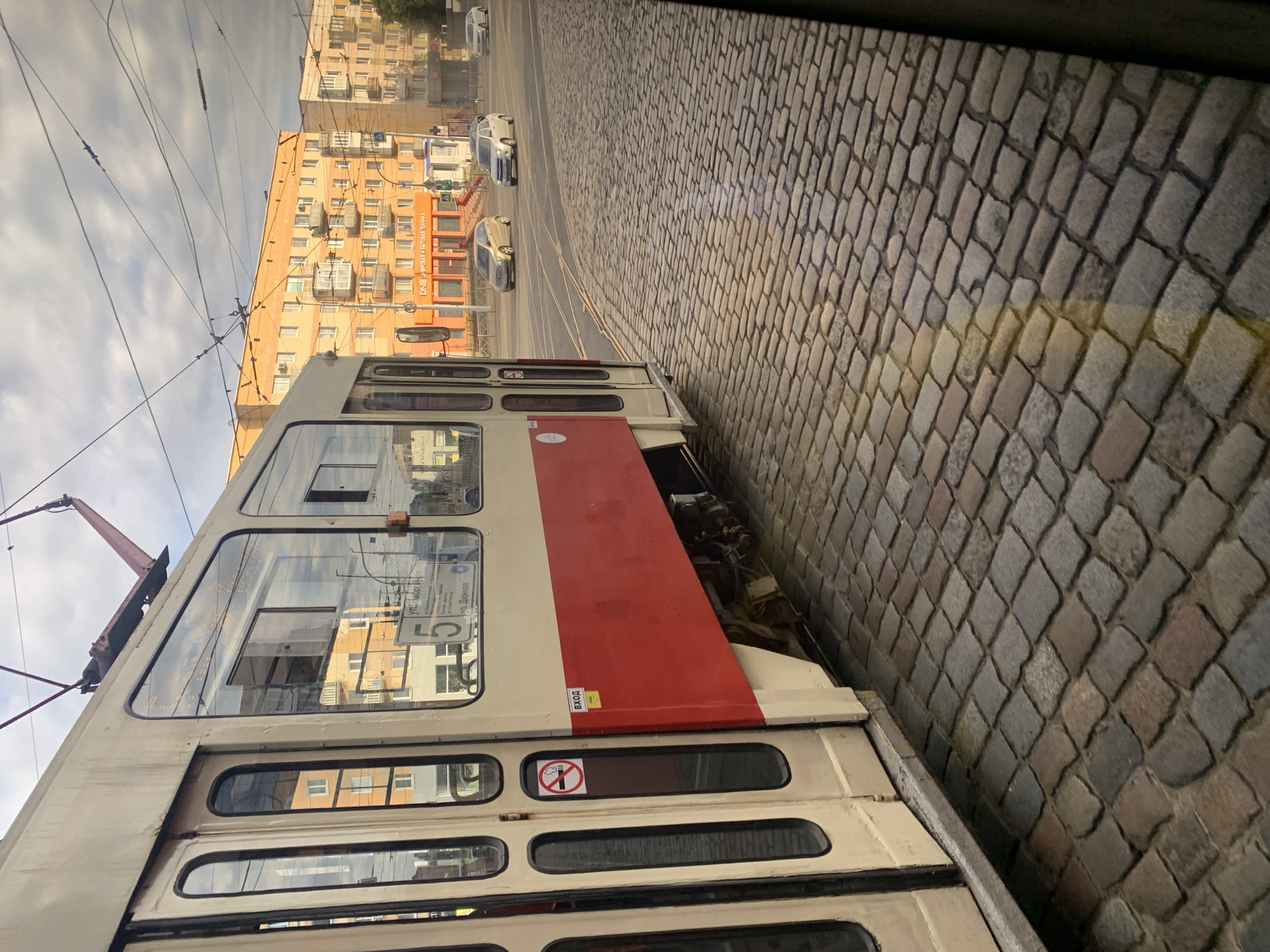 A tram is stopped at a street corner, with its doors open to allow passengers to board and alight. The tram is cream and red, and it is on a cobblestone street. In the background, there are buildings with balconies and windows, and a few cars are driving on the street. The sky is cloudy. A "no smoking" sign is visible on the tram door, and a yellow sign with "ВХОД" (entrance) is also visible. The tram appears to be on route number 5, indicated by a sign on the side.