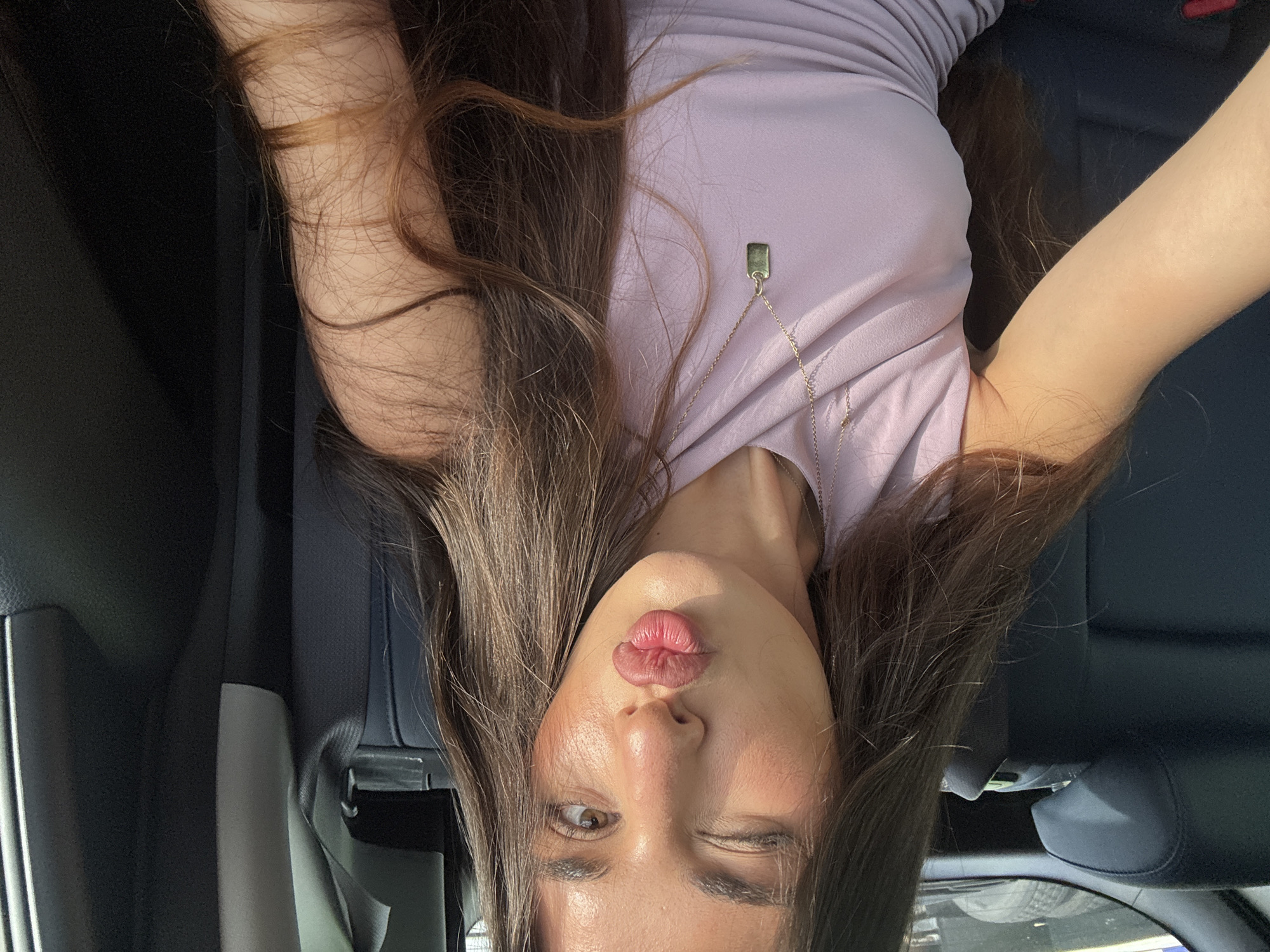A woman is inside a car, possibly the backseat. She is wearing a light purple top and a necklace with a pendant. She is winking with her right eye and puckering her lips. The car has dark blue leather or leatherette seats and interior trim. The image orientation is upside down. The location is unspecified, but the scene is in Boca Raton, United States of America.
