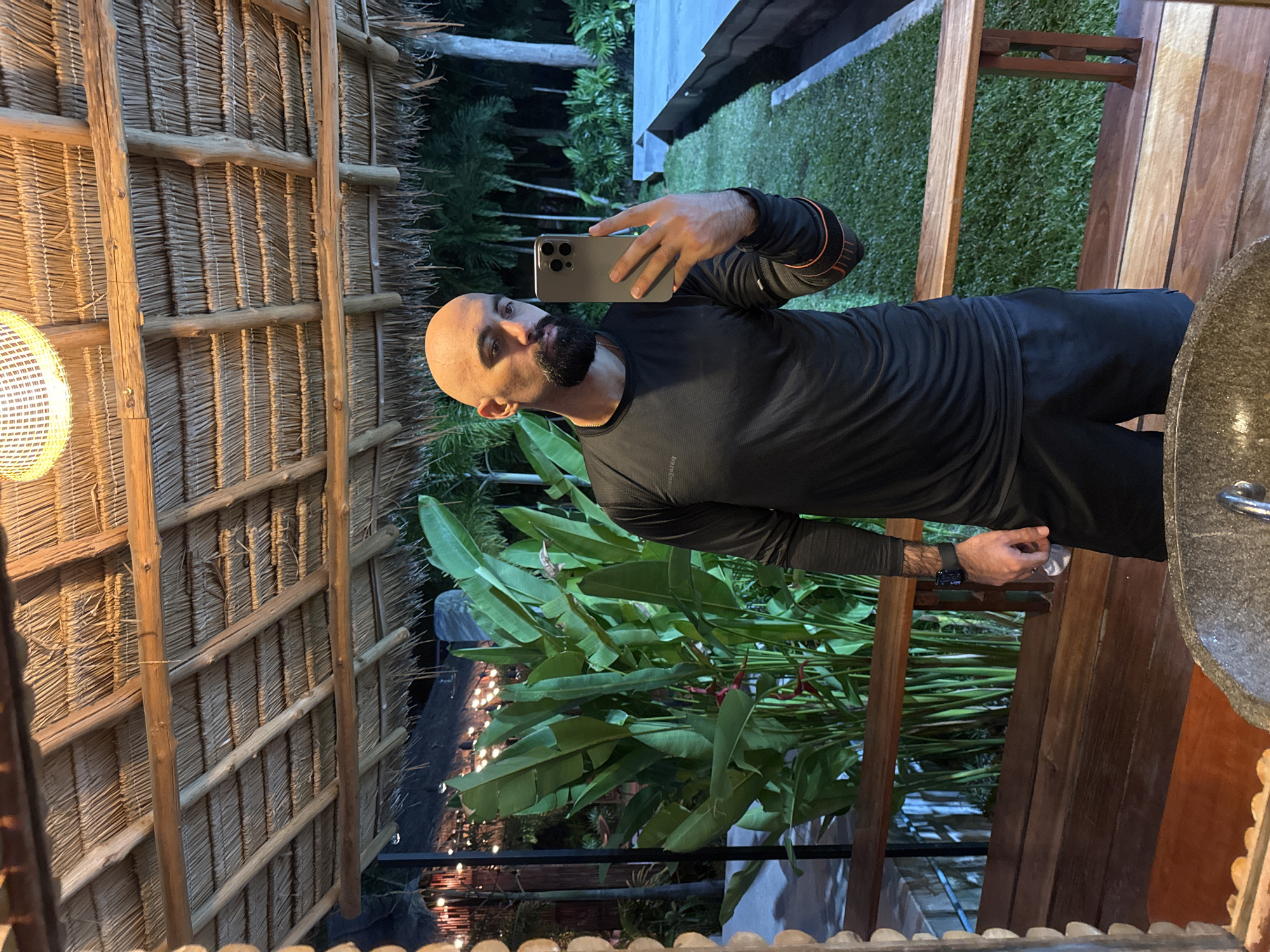 A bald man with a beard is taking a selfie in a tropical setting. He is wearing a black long-sleeved shirt and black shorts. The background shows lush green foliage, palm trees, and a grassy area. The man is holding a silver smartphone in his right hand. The scene appears to be outdoors, possibly on a patio or balcony, with a thatched roof overhead and wooden structures. The lighting suggests it is either dusk or nighttime. The image does not contain any recognizable landmarks that would pinpoint the specific location within Ko Samui District, Thailand.