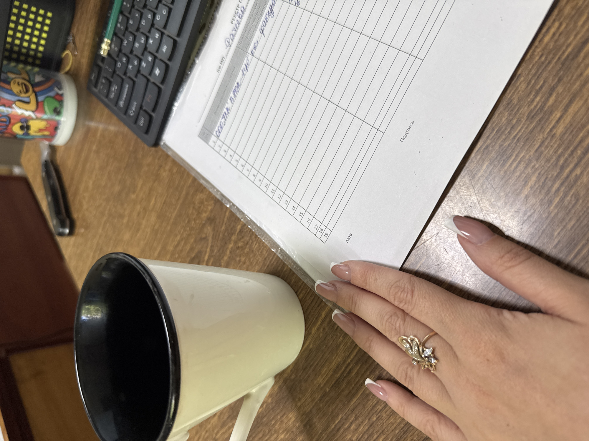 The image shows a close-up, high-angle shot of a desk with a mug, a hand with a ring, and a document with handwritten text. The desk is made of wood. A white mug with a black interior sits on the left side of the frame. A woman's hand with manicured nails and a gold ring adorned with small stones is positioned over the document. The document appears to be a form with columns and rows, and it contains handwritten Cyrillic text. A black keyboard is visible behind the document, and a small, colorful mug with cartoon characters is in the background. A pen is resting on the keyboard. The scene suggests a workspace environment.