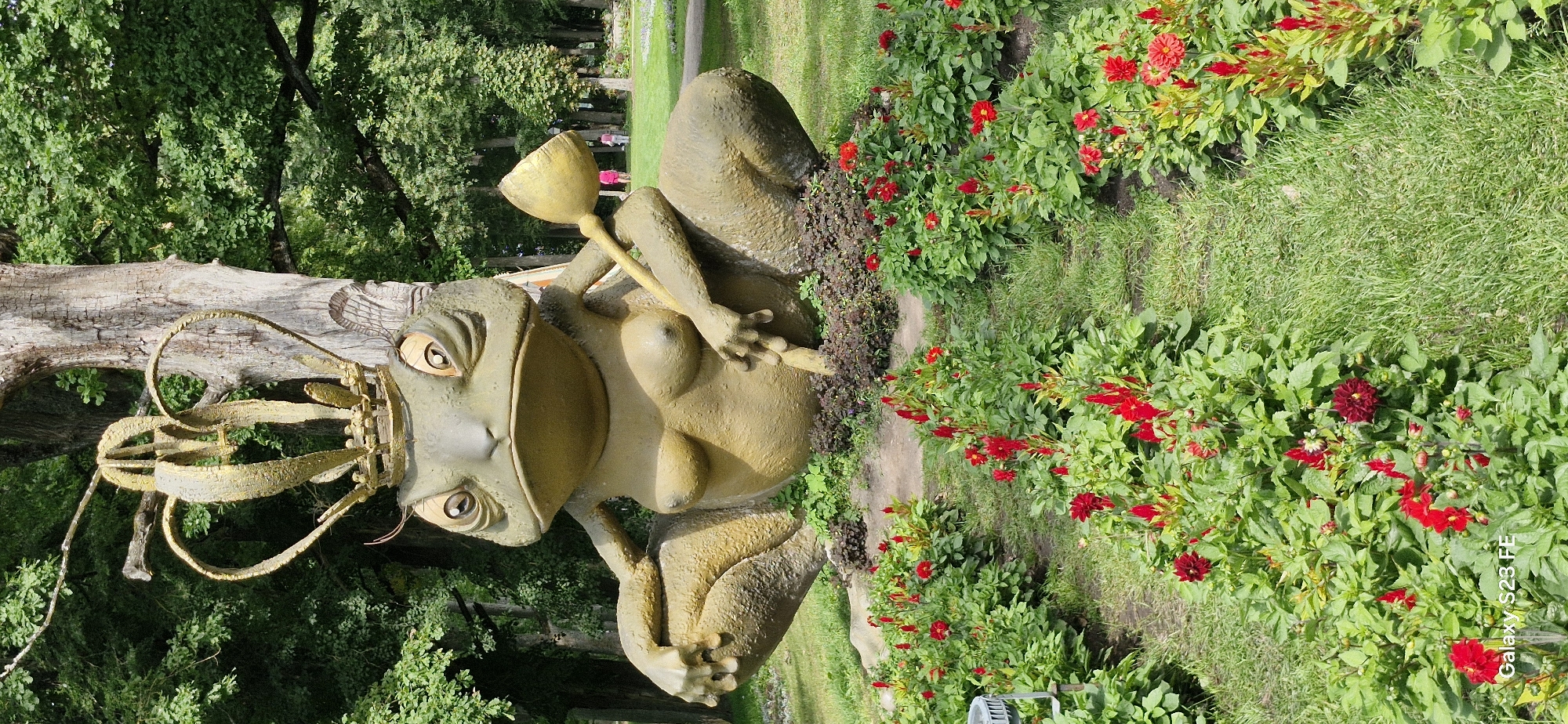 The image captures a large, weathered sculpture of a crowned frog, resembling a queen, seated on a grassy mound. The frog statue is green and appears to be made of metal or concrete, with a golden crown adorned with heart shapes on its head. It is holding a golden scepter-like object in its right hand. The frog statue is positioned in the foreground, surrounded by a bed of vibrant red flowers and green foliage. In the background, a large tree stands behind the sculpture, and beyond that, a park-like setting with more trees, green grass, and a pathway is visible. Two individuals, appearing as children or young adults, are walking in the distance on the pathway. The setting is outdoors, suggesting a park or garden area in Pakruojis, Lithuania. The overall impression is of a whimsical and somewhat unusual public art installation within a natural environment.