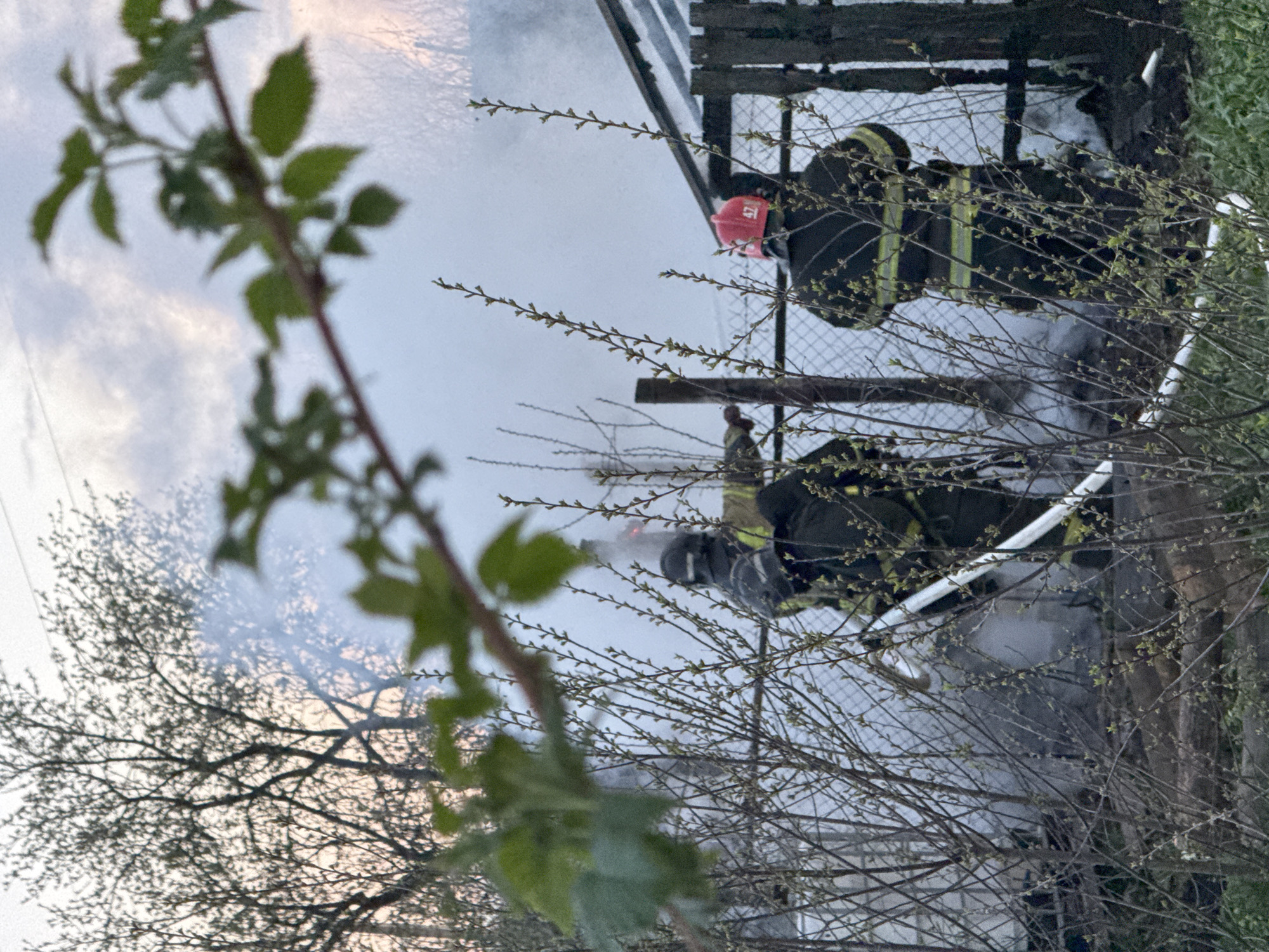 The image, rotated counterclockwise, depicts firefighters responding to an incident in Chaikovsky, Russia. Two firefighters are visible, wearing dark uniforms with yellow reflective stripes and helmets. One firefighter has a red helmet and is standing above the other. Both firefighters are positioned near a fence, partially obscured by branches in the foreground. A large amount of smoke is present, partially obscuring the building in the background. The sky is overcast. Trees and branches are in the foreground and background.