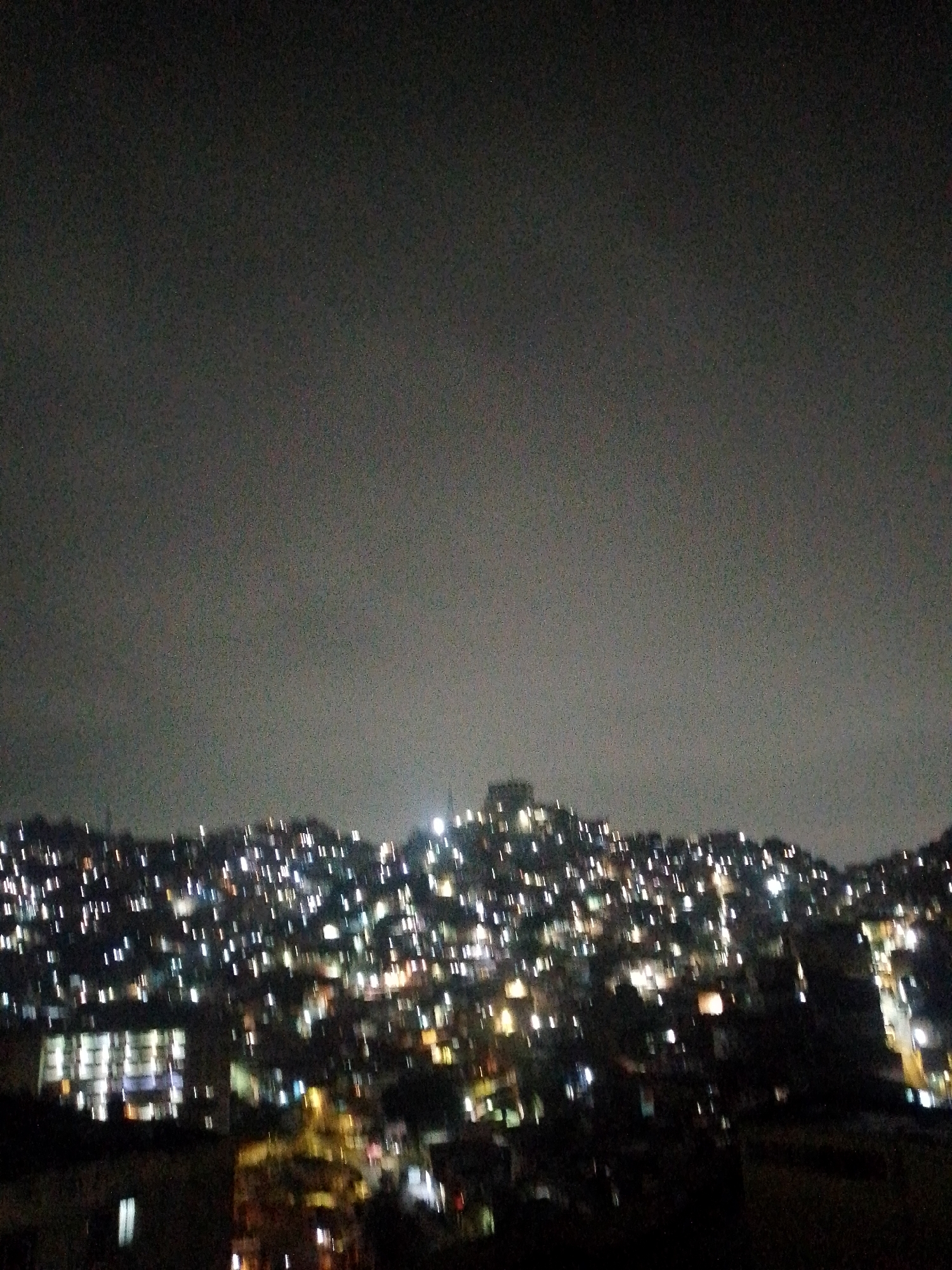 A night view in Caracas, Venezuela, shows a densely populated hillside covered with numerous lights from residences. A tall structure with lit windows stands out in the foreground. The sky is overcast and uniformly gray.