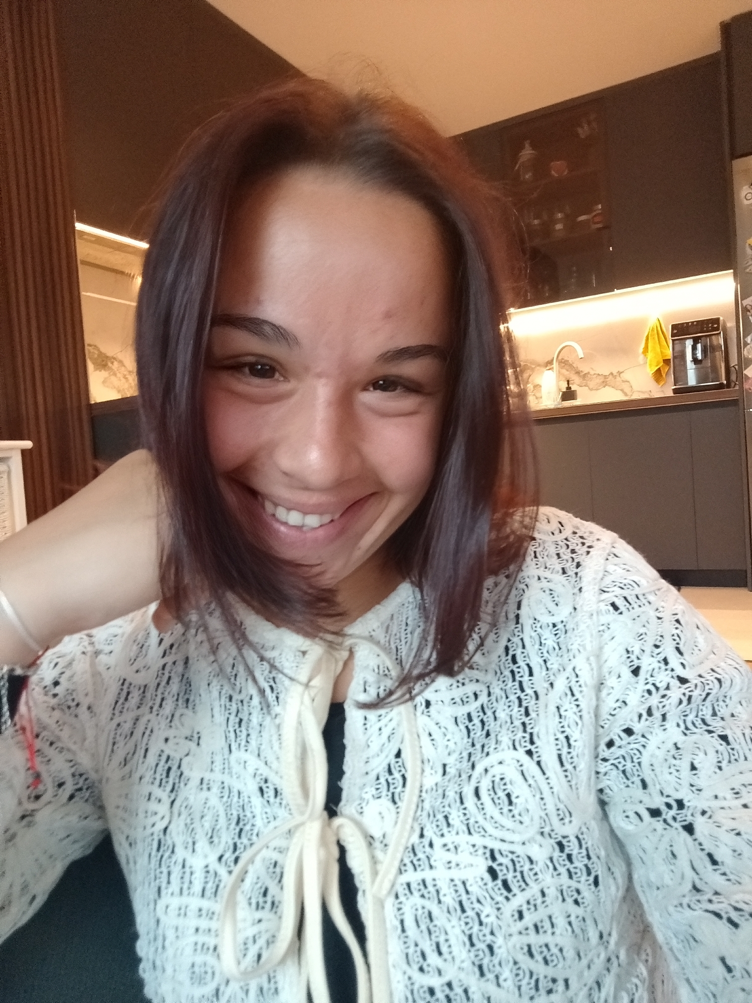An eye-level, medium close-up portrait shows a young woman smiling broadly indoors, likely in a modern home in Sofia, Bulgaria. She has medium-length, dark brown hair, dark eyes, and a radiant smile revealing her teeth, with her cheeks creased in dimples. Her head is tilted slightly, and her left hand is casually resting near her chin, with a thin, light-colored bracelet visible on her wrist.
She is wearing a cream or white open-knit garment, possibly a cardigan or jacket, featuring intricate patterns and tied at the front with light-colored strings. A dark-colored top is visible underneath.
The background reveals a contemporary interior. To the left, a vertical slatted wooden wall or partition is partially visible, alongside a glimpse of light-colored furniture. To the right, a modern kitchen area features dark grey, minimalist cabinets. A light-colored, patterned countertop (resembling marble or granite) holds a white gooseneck faucet, a black soap dispenser, and a black coffee machine. Under-cabinet strip lighting casts a warm glow on the backsplash. A bright yellow towel hangs on the wall near the coffee machine. Above the counter, dark-framed glass-front cabinets display various items. The lighting in the room is bright, suggesting daytime. The woman's expression conveys happiness and cheerfulness. No discernible text is visible in the image.