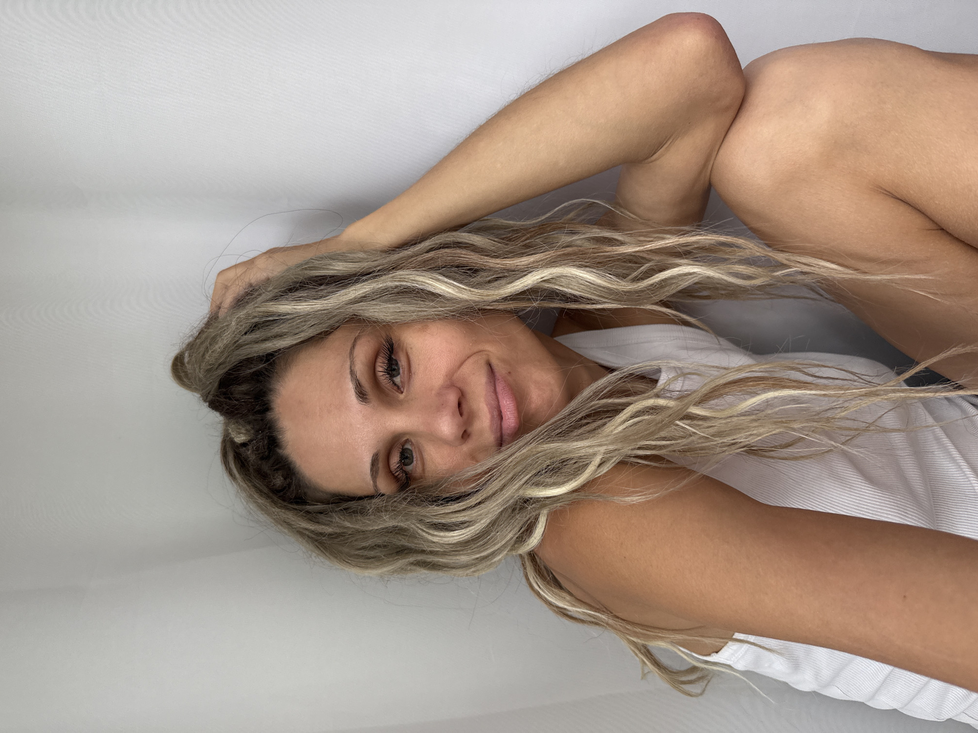 A blonde woman with long, wavy hair is shown in a seated pose. She is wearing a white tank top and has her right arm raised, with her hand resting on her head. The background is a plain, light-colored wall. The scene appears to be indoors, possibly a studio or a room. There is no indication of the location being in Rusanovo, Russia.