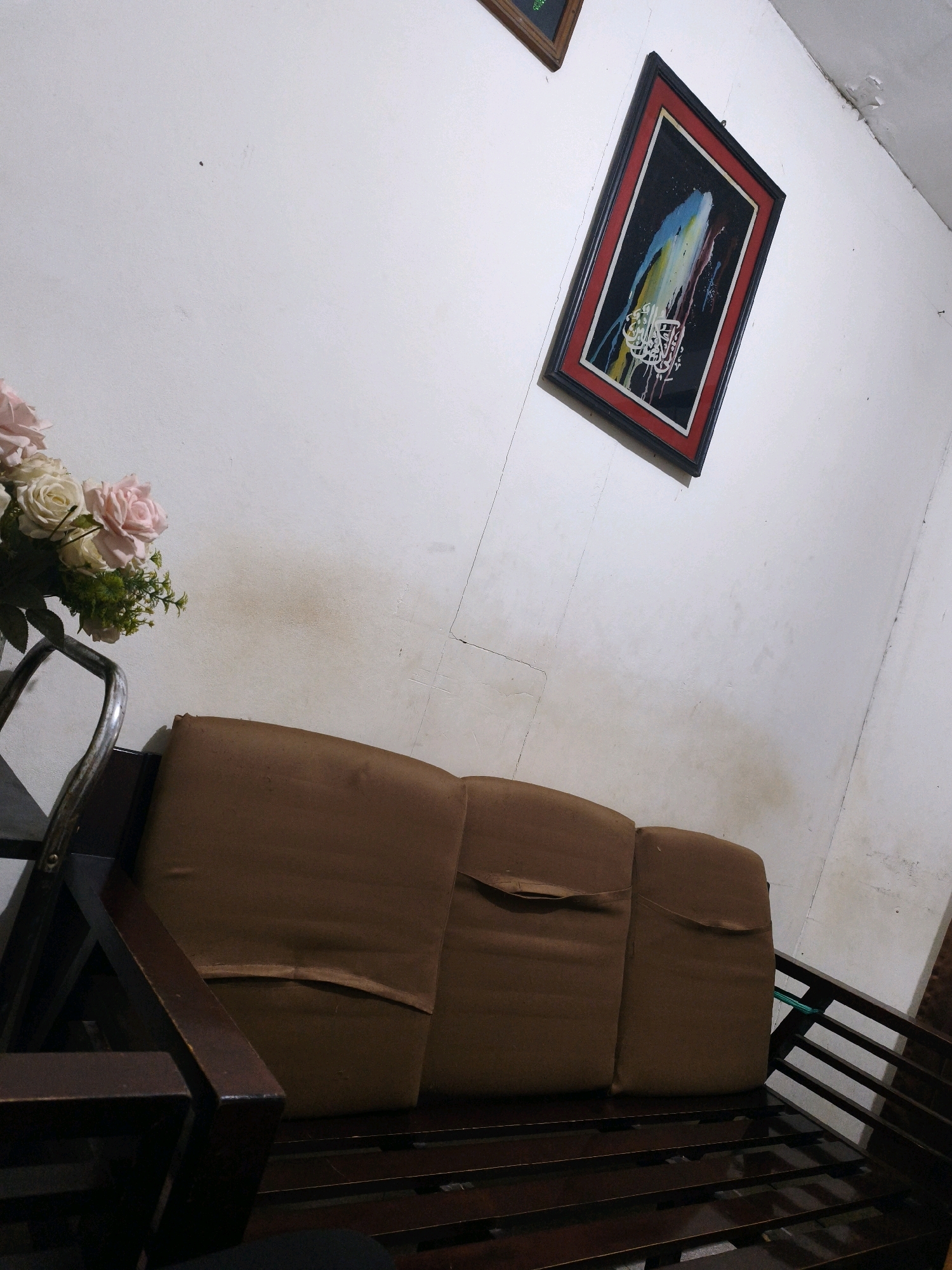 An interior shot captures a dimly lit room in Samarinda, Indonesia. The focal point is a dark wooden sofa with three brown cushions, showing signs of wear. To the left, a bouquet of artificial pink and white roses sits in a vase, partially obscured by the sofa's armrest. Above and to the right, a framed picture hangs on a white wall; the frame is black with a red inner border, and the artwork within depicts abstract colorful strokes over Arabic calligraphy. The wall exhibits some discoloration and minor damage. The overall scene suggests a lived-in residential space.