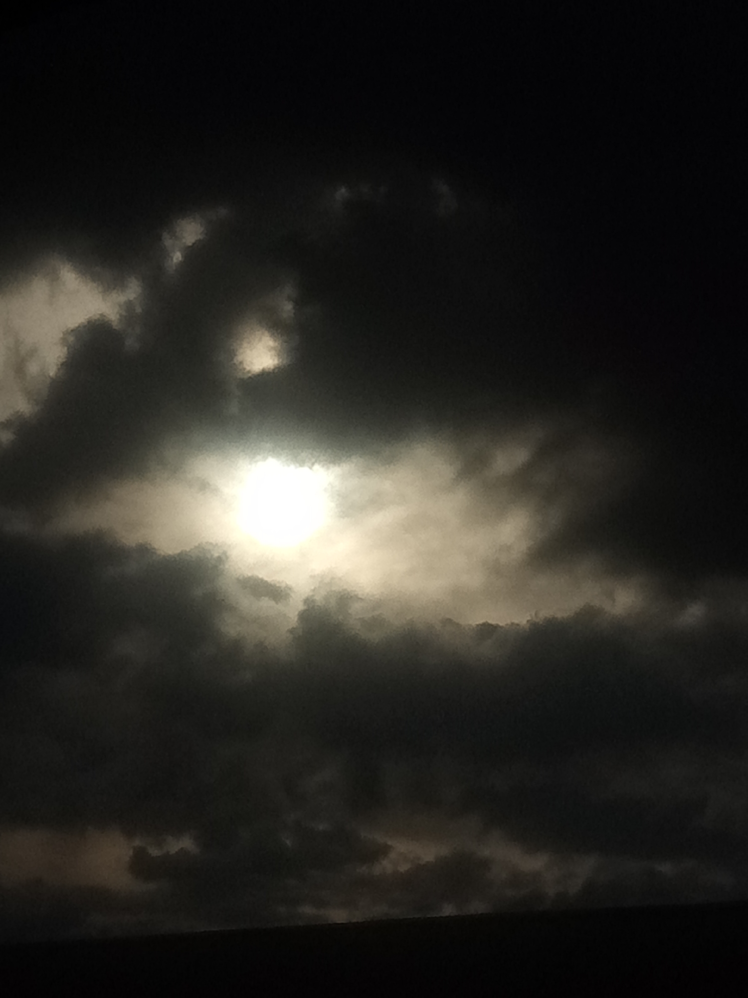 The image is a nighttime photograph showing a bright, full moon partially obscured by dark, textured clouds. The moon's light creates a luminous glow that diffuses through the thinner parts of the clouds, casting a soft, ethereal light on the scene. The clouds are dense and varied in shape, creating a dramatic and somewhat ominous atmosphere. The overall impression is one of a celestial event taking place in the dark of night. The photograph is taken from a low angle, with a dark silhouetted landscape at the bottom of the frame, suggesting a view from the ground looking upwards.