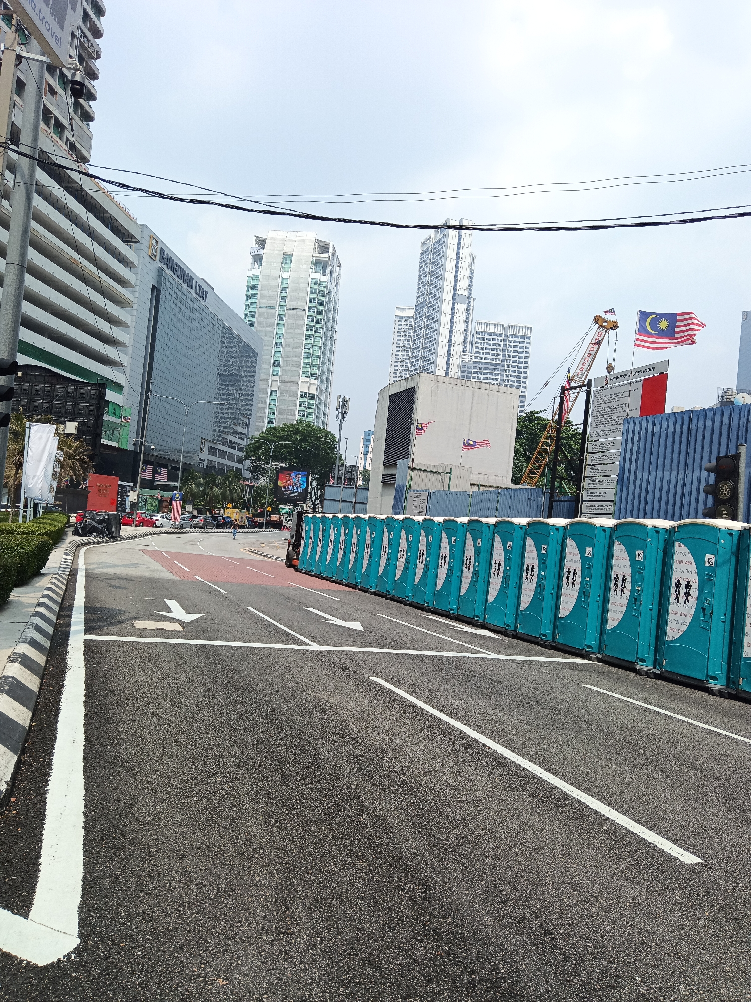 The image captures a street scene in Kuala Lumpur, Malaysia, with a line of teal portable toilets on the right side of the road. The toilets are arranged in a row and have black and white silhouettes of people on them. The road itself is asphalt with white lane markings and arrows indicating direction. In the background, several tall buildings are visible, including a modern glass-fronted skyscraper with the name "BANGUNAN LTAT" prominently displayed. A construction crane is also present, suggesting ongoing development. The Malaysian flag is flying from a flagpole. The sky is overcast, and the overall environment appears to be an urban setting with a mix of infrastructure and development. There are a few cars visible on the road, and a single person is walking in the distance. The scene is static, with no obvious actions or interactions taking place.