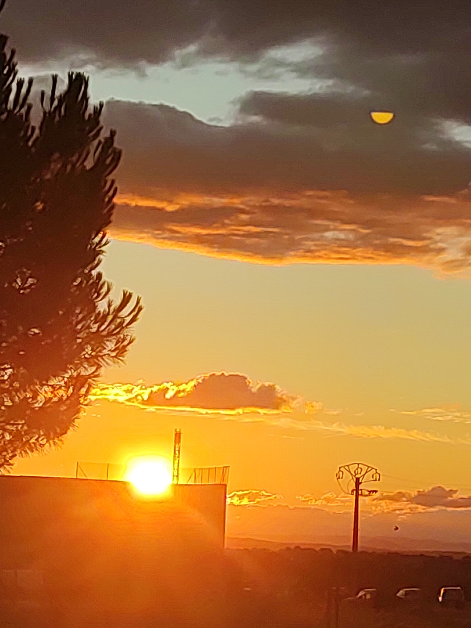 This is a vertical, medium-shot photograph taken at sunset in Sobradillo, Spain. The scene depicts a vibrant orange sky filled with dramatic clouds, which are illuminated by the setting sun. In the foreground, a dark silhouette of a pine tree is visible on the left side. A building, also in silhouette, runs across the bottom half of the image, with the sun appearing as a blinding white orb just above its roofline. A thin tower or antenna rises from the roof. To the right, a utility pole with multiple crossbars stands in the middle ground, with a distant mountain range visible on the horizon. A small, crescent-shaped object, possibly the moon, is faintly visible in the upper portion of the sky, amidst dark clouds. The overall atmosphere is one of intense color and dramatic natural light.