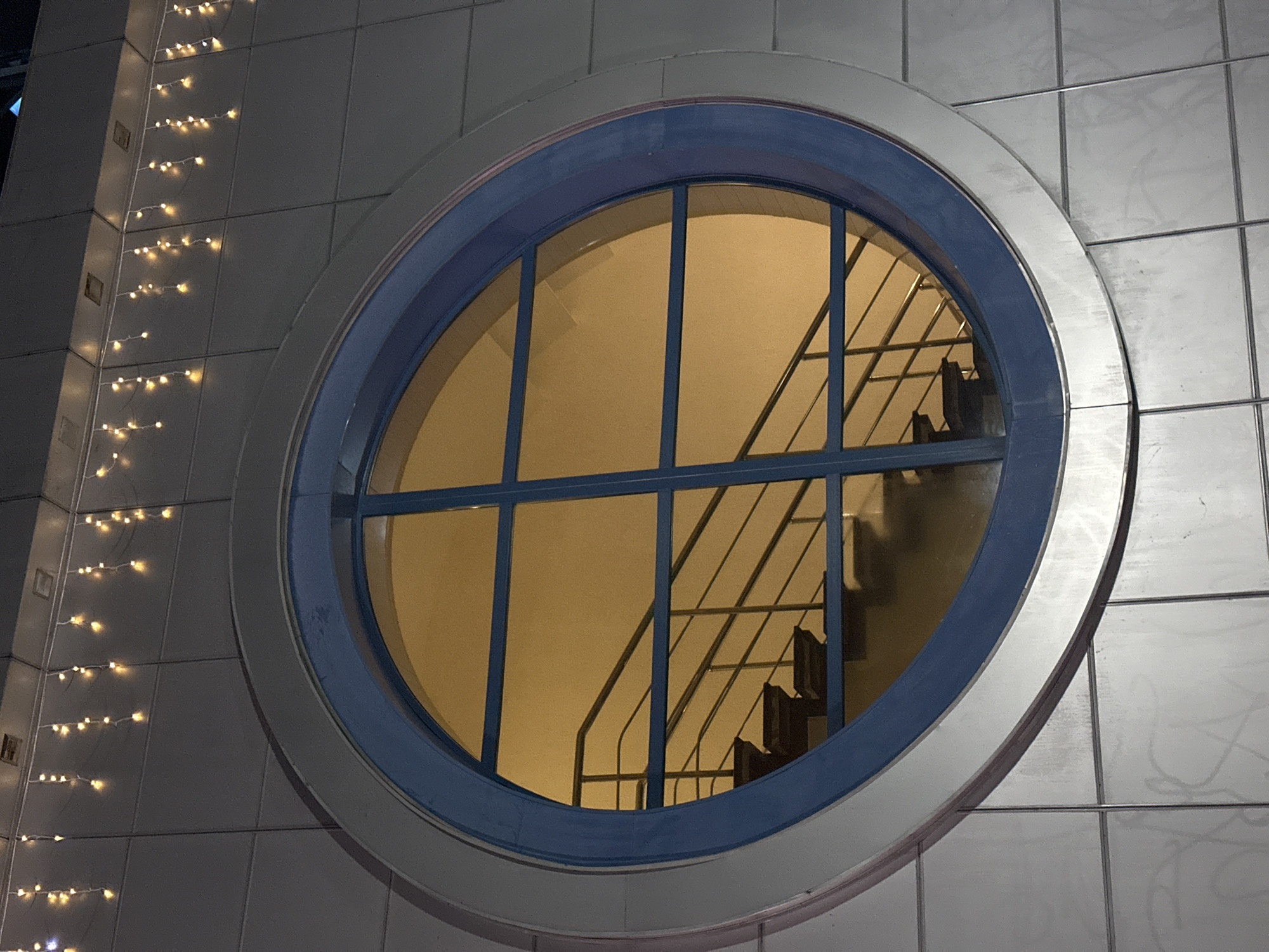 This is a medium shot, low-angle, exterior photograph of a large, circular window on a modern building. The window has a blue frame and is divided into four sections by a dark blue grid. The glass is reflecting warm, yellow light, and through it, a dark staircase with a metal railing is visible, ascending into darkness. Above the window, on the grey tiled facade of the building, a string of small, yellow lights is draped, resembling icicle lights. The building exterior appears to be constructed of large, square tiles, some of which have faint, grey markings on them. The photograph was taken at night or in dim light conditions. The location is the city of Tomsk, Russia. There are no people or animals visible in the image.