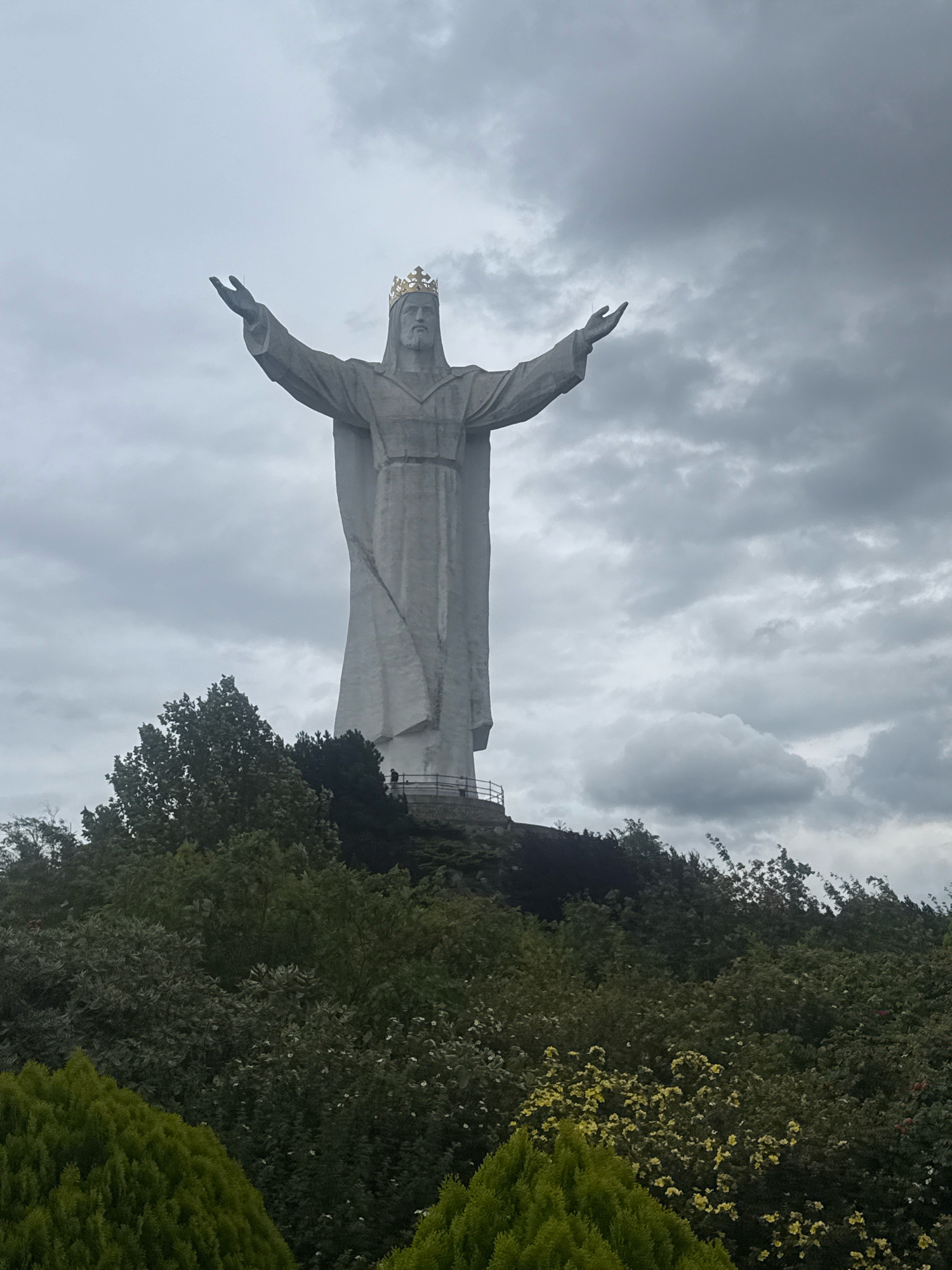 A large, imposing white statue of Jesus Christ stands atop a hill, its arms outstretched against a cloudy, overcast sky. The statue is adorned with a golden crown. Lush green trees and foliage surround the base of the hill, creating a stark contrast with the pale figure of the statue. The statue is located in the city of Świebodzin, Poland, and is known as the Christ the King statue.