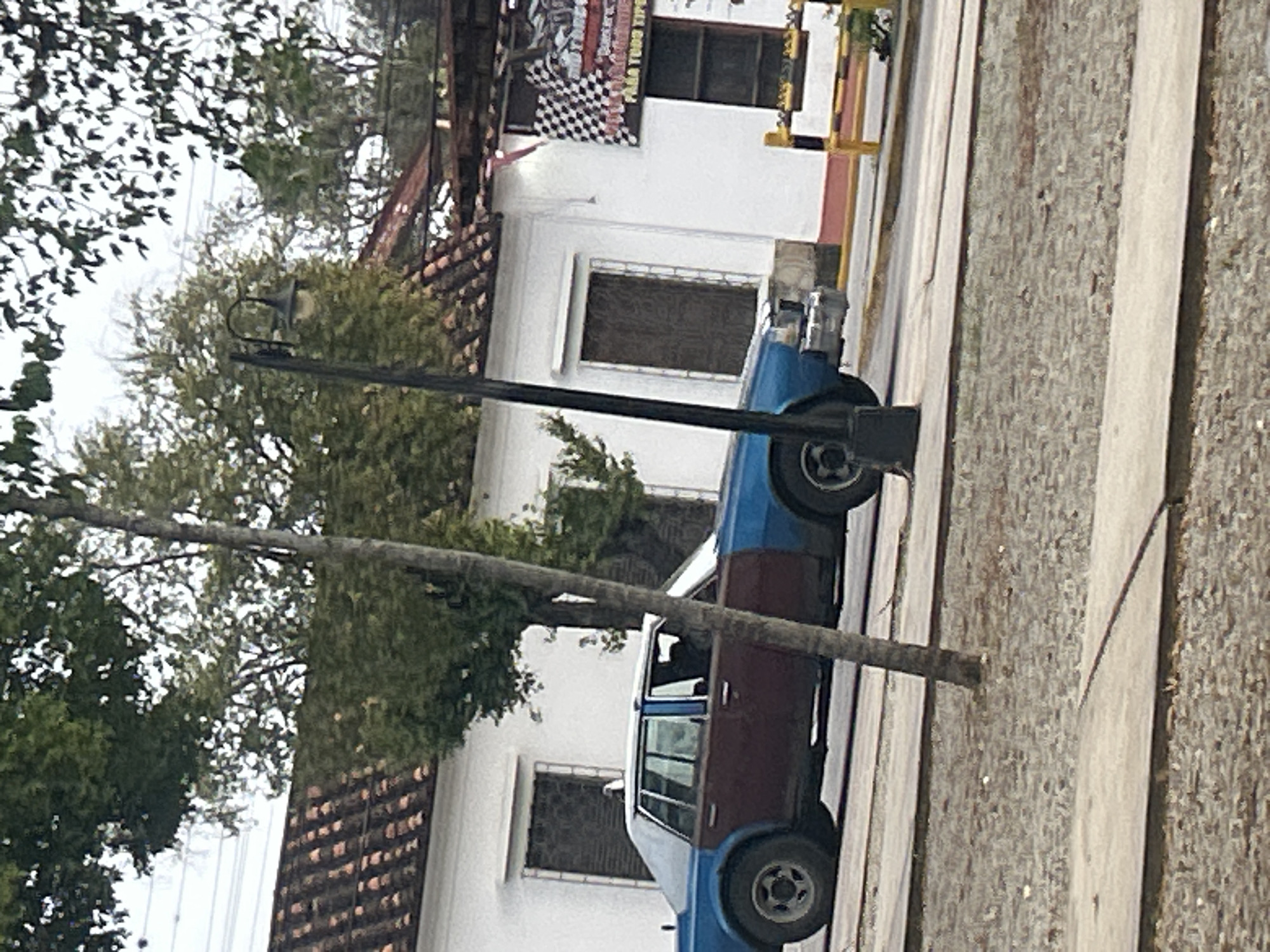 The image depicts a two-toned sedan, with the front portion painted blue and the rear section in a dark reddish-brown color, parked on a street in Santa Ana, Venezuela. The car has tinted windows. To the left of the car, a slender tree trunk is visible, and further left, a white building with a tiled roof and dark-grated windows is partially obscured by foliage. On the right side of the image, another white building is visible, featuring a checkered flag banner and a yellow barrier. A lamppost stands in front of the car. The ground in the foreground consists of a paved walkway and a gravelly area. The overall scene suggests an outdoor urban environment.