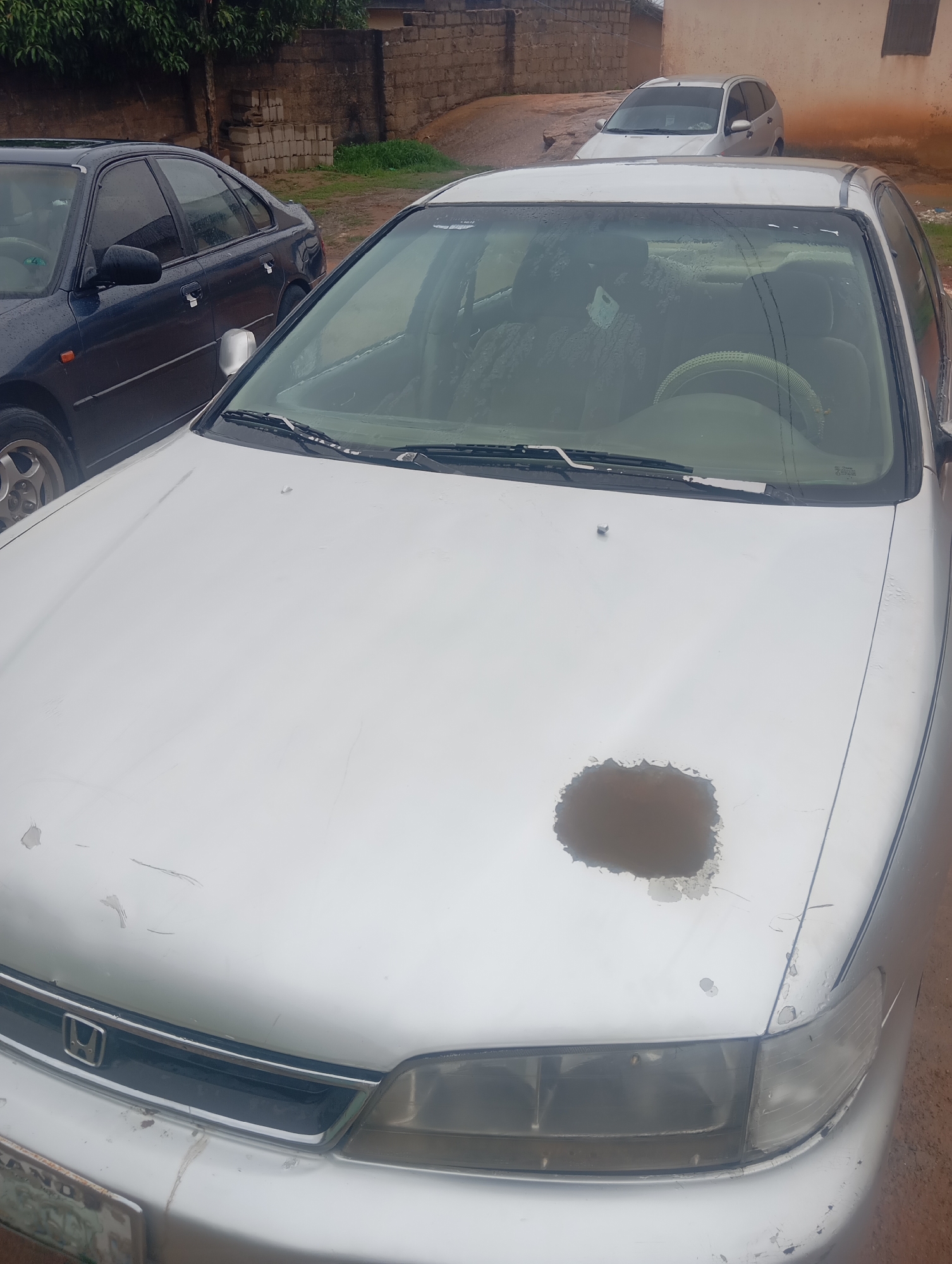 The image shows a silver sedan parked on a dirt road in front of a brick wall. The car's hood has a large patch of paint damage. In the background, another silver car is parked. The environment appears to be an outdoor urban setting, possibly in Jos, Nigeria. The lighting suggests it is daytime, and the ground is wet, indicating recent rain. There are no people visible in the image. The overall scene depicts a street-level view of parked vehicles in a modest urban environment.