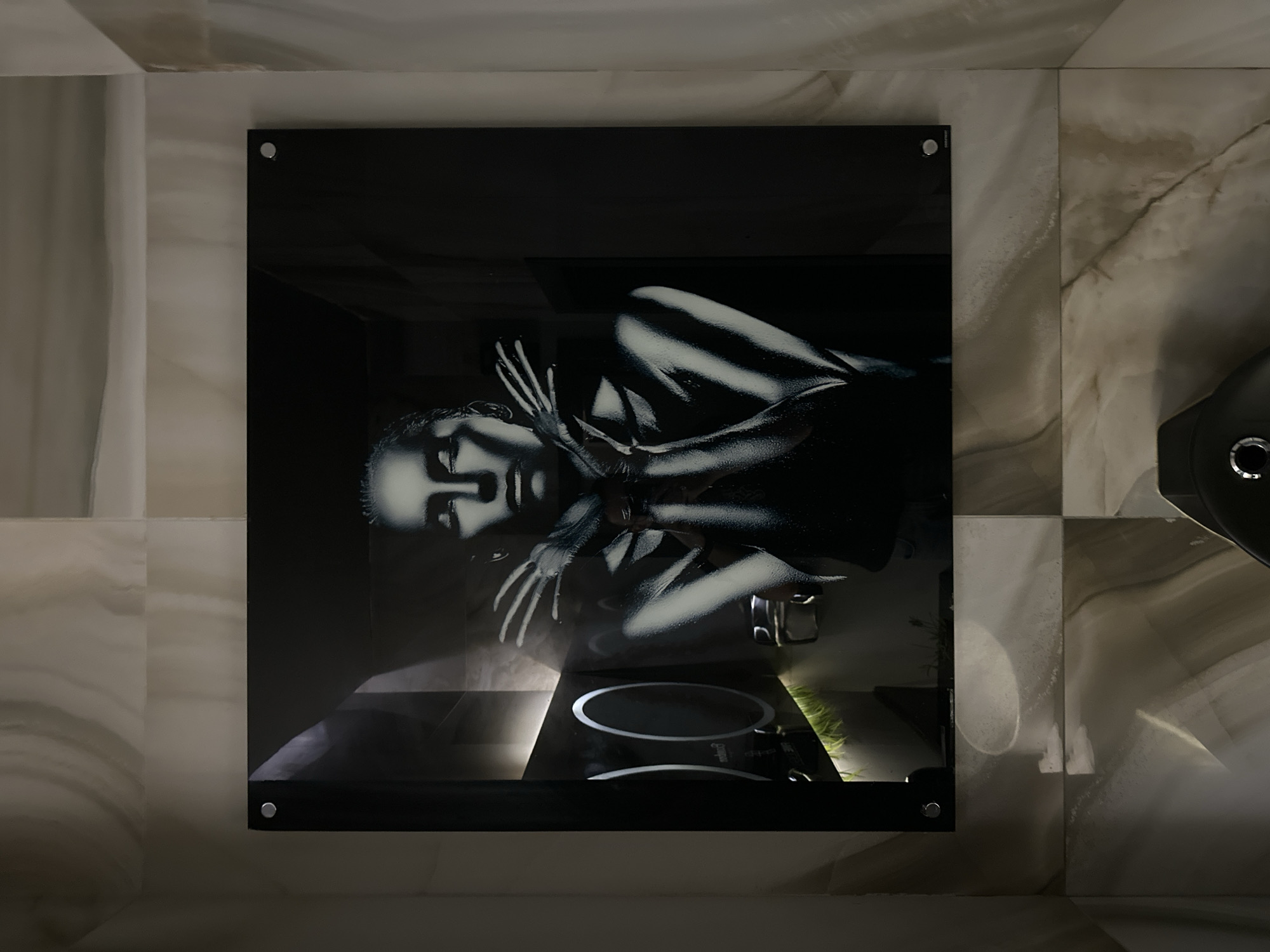 The image shows a framed black and white artwork depicting a woman lying down with her hands near her face. The artwork is mounted on a marble wall. Below the artwork, there appears to be a black stovetop. The location is not discernible from the image. The scene presents an artwork within a bathroom setting.