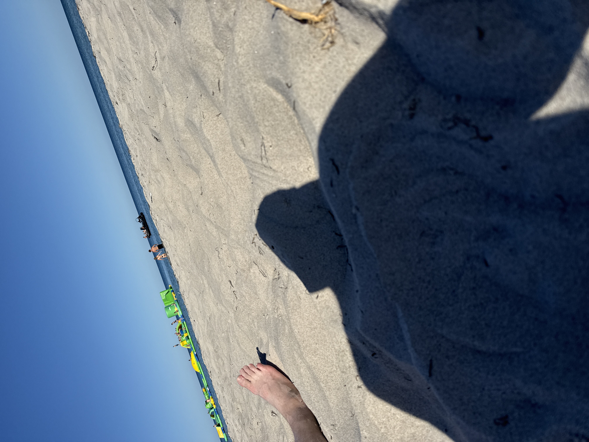 The image depicts a sandy beach with the ocean meeting the shore. The composition is at an angle, with the sand occupying the majority of the frame, and the clear blue sky and ocean forming a thin strip on the left. In the foreground, a bare foot is visible, casting a shadow on the sand. Further down the beach, several people are seen in and near the water. Some are standing, while others are lying on the sand. There is also an inflatable water park structure in bright green and yellow, with people interacting with it. The sunlight suggests it is daytime, likely a sunny day given the clear sky and shadows. The scene evokes a sense of leisure and recreation at a beach resort.