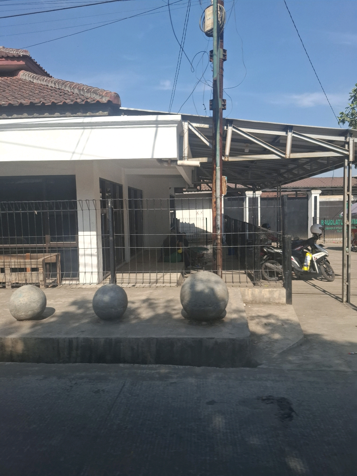 The image depicts a street scene in Cisaat, Indonesia, under a clear blue sky. In the foreground, a concrete curb separates the asphalt road from a raised concrete platform. On this platform sit three large, spherical concrete bollards, casting shadows on the ground.
Behind the bollards is a building with a white facade and a red-tiled roof. A metal fence runs along the front of the building, with a gap where a black metal gate is visible. Through the fence, a wooden table and chair can be seen. To the right, a covered carport area is visible, supported by metal beams. Under the carport, two motorcycles are parked. One motorcycle is white with black accents and a yellow water bottle attached to its side. The other motorcycle is dark-colored. A helmet rests on the seat of the white motorcycle.
A utility pole with numerous wires is positioned to the left of the carport structure, with a meter box attached to it. Further to the right, a building with a sign that reads "RAUDLATUL ATFAI..." is partially visible. The overall environment suggests a commercial or residential area with some public access.