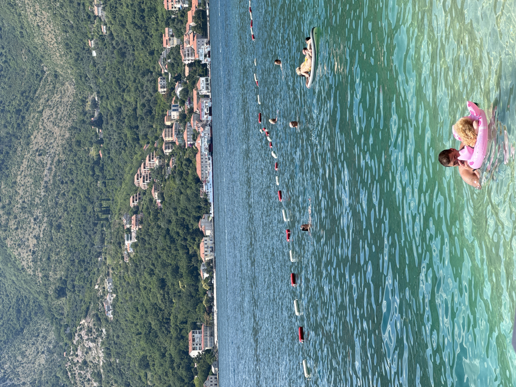 This media file depicts a scenic view of a coastal town nestled against a lush green mountainside, with a clear, turquoise body of water in the foreground. The buildings, predominantly white with red-tiled roofs, are scattered along the slopes, indicating a Mediterranean or similar climate. The water is calm and inviting, with a line of red and white buoys marking a designated swimming area.
In the water, several people are enjoying the day. Two individuals are on an inflatable raft, appearing to be relaxing or sunbathing. Closer to the viewer, a man is holding a young child who is wearing a pink inflatable ring. They are both in the water, suggesting a playful or caring interaction. The water is transparent enough to see the sunlight shimmering on the ripples and the sandy bottom in shallower areas.
The scene suggests a warm, sunny day, likely during daylight hours given the clear visibility and bright lighting. The overall atmosphere is one of leisure and enjoyment. The juxtaposition of the built environment with the natural landscape of mountains and sea creates a picturesque setting. There is no visible text in the image.