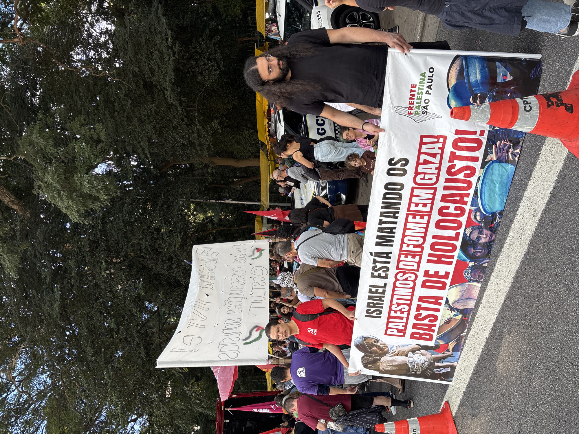 The image shows a protest with people holding banners in what appears to be a street in São Paulo, Brazil. One banner reads "ISRAEL ESTÁ MATANDO OS PALESTINOS DE FOME EM GAZA! BASTA DE HOLOCAUSTO!" ("ISRAEL IS KILLING PALESTINIANS OF HUNGER IN GAZA! ENOUGH OF HOLOCAUST!"). Another banner reads "FRENTE PALESTINA SÃO PAULO". The protest is surrounded by trees and there are traffic cones nearby. Several people are visible holding the banners and participating in the protest. A police car is also visible.
