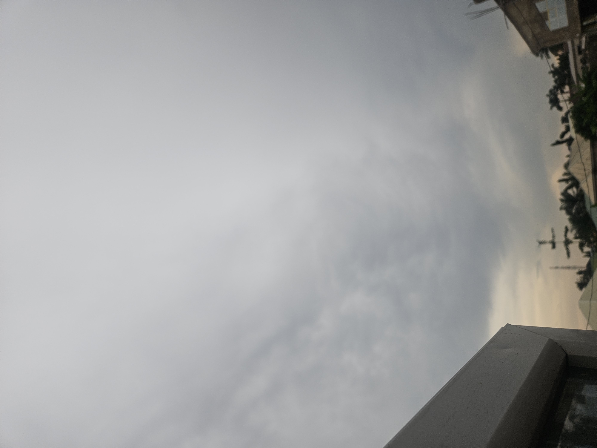The image shows an overcast sky above a cityscape, with the foreground dominated by the corner of a white building structure. In the background, several buildings with varied rooflines, trees, and a communication tower are visible. The sky is filled with dark, heavy clouds, suggesting an impending storm or a generally gloomy weather condition. The scene appears to be from an urban or semi-urban environment in Masaka, Nigeria.