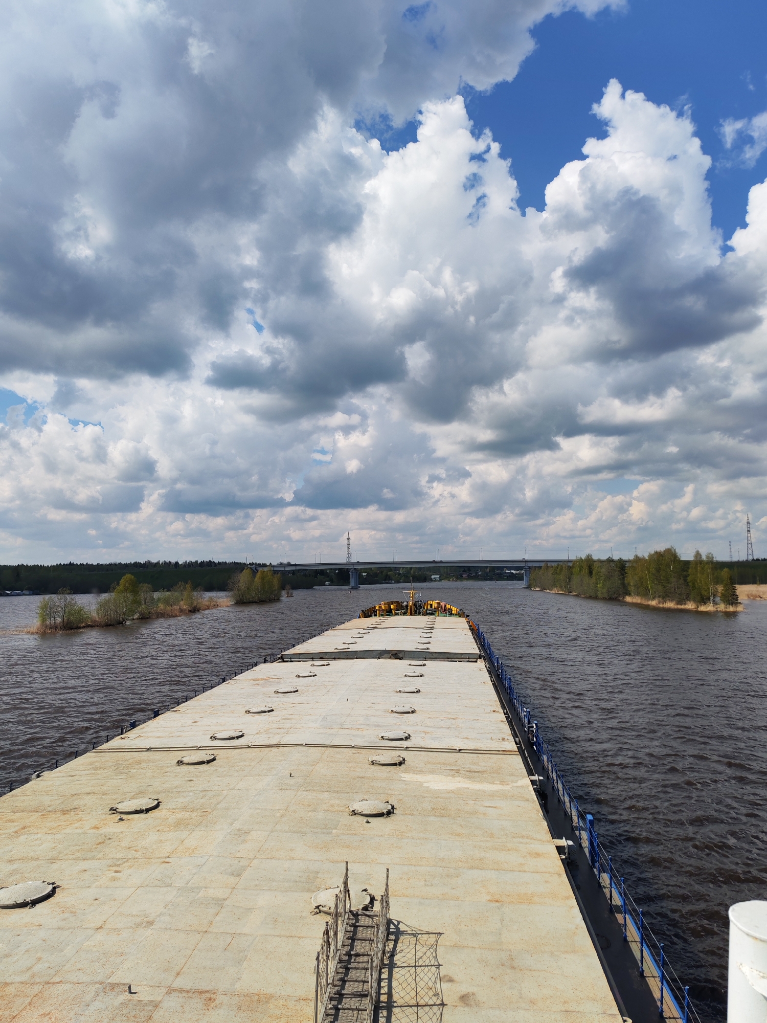 The image depicts a wide, flat barge moving on a waterway, presumably a river or wide canal. The surface of the barge is largely featureless, with evenly spaced, circular access hatches. A blue railing runs along the edge of the barge. The water on either side is dark and choppy. Along the banks, there are trees and shrubs, with patches of what appears to be marshy vegetation. In the distance, a bridge spans the waterway, supported by pylons. The sky is filled with white and gray clouds, with patches of blue visible. The location is unknown, Unknown.