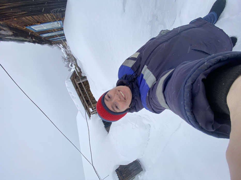 A female subject is depicted in a selfie, smiling directly at the camera in a heavily snow-covered outdoor environment in Узяр, Russia. She wears a red and blue knitted beanie and a dark blue winter parka with a royal blue upper section and grey reflective bands on the sleeves. Her right arm is extended, holding the camera, with a gloved hand partially visible in the foreground. The ground is covered in deep snowdrifts, which also blanket roofs and obscure lower portions of structures.
In the background, to the right, a weathered log house is visible, featuring three windows with light blue wooden shutters; accumulated snow reaches approximately window-sill height. Centrally, a wooden lean-to structure, containing neatly stacked firewood, stands with its roof heavily covered in snow. To the far left, a small, dark wooden outbuilding is partially submerged in snow. A single black utility line traverses the upper third of the frame. The sky is uniformly overcast, indicating a winter day.