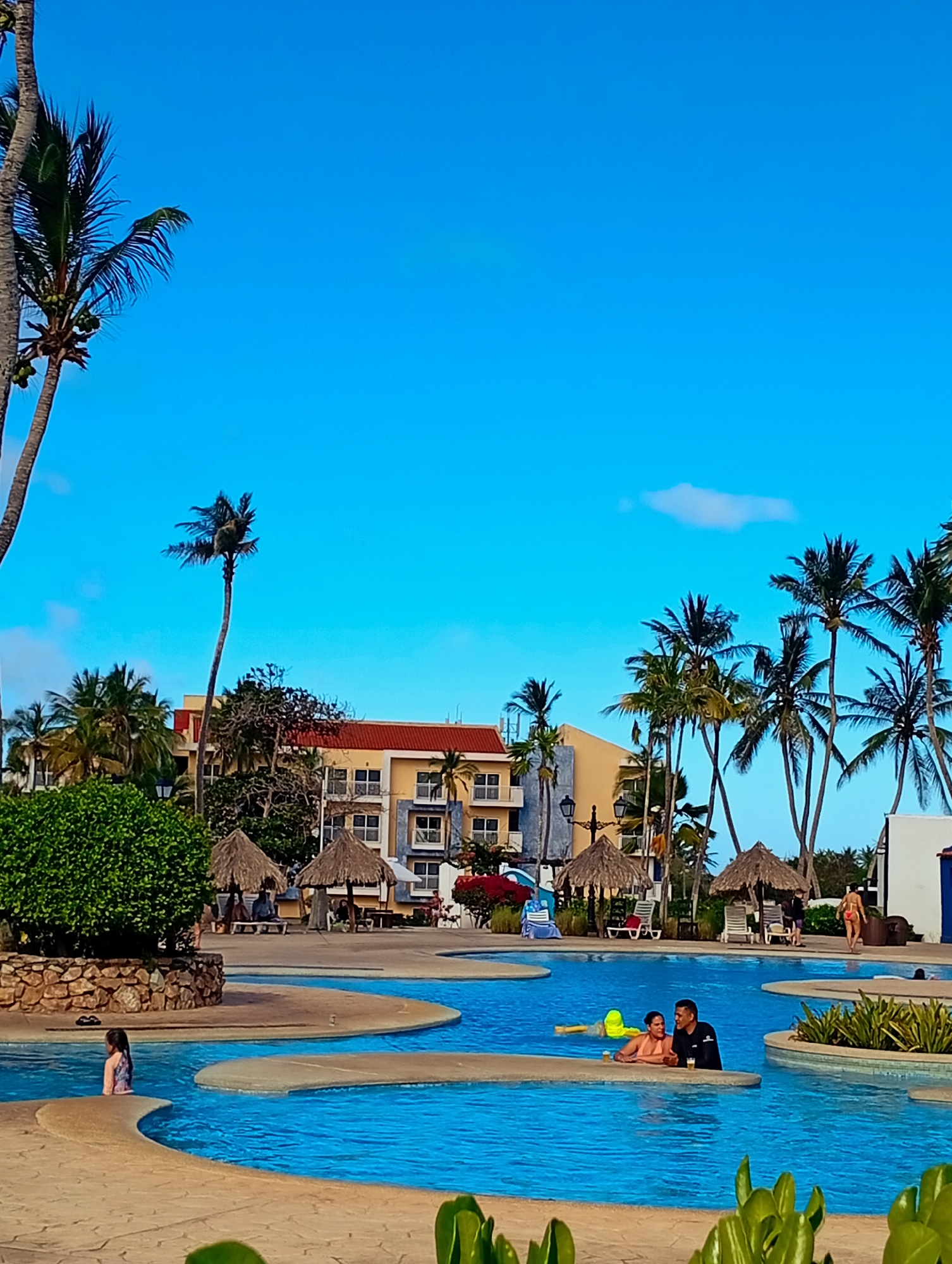 The image depicts a scene at a resort in Playa El Agua, Venezuela. A multi-tiered swimming pool occupies the foreground, filled with bright blue water. In the pool, a child and an adult couple are visible.
The resort area features several palm trees and thatched-roof cabanas. In the background, a building with yellow walls and a red tiled roof is visible. The sky above is clear blue with a few scattered clouds. The scene suggests a tropical vacation setting with people enjoying leisure activities in a resort-style environment.