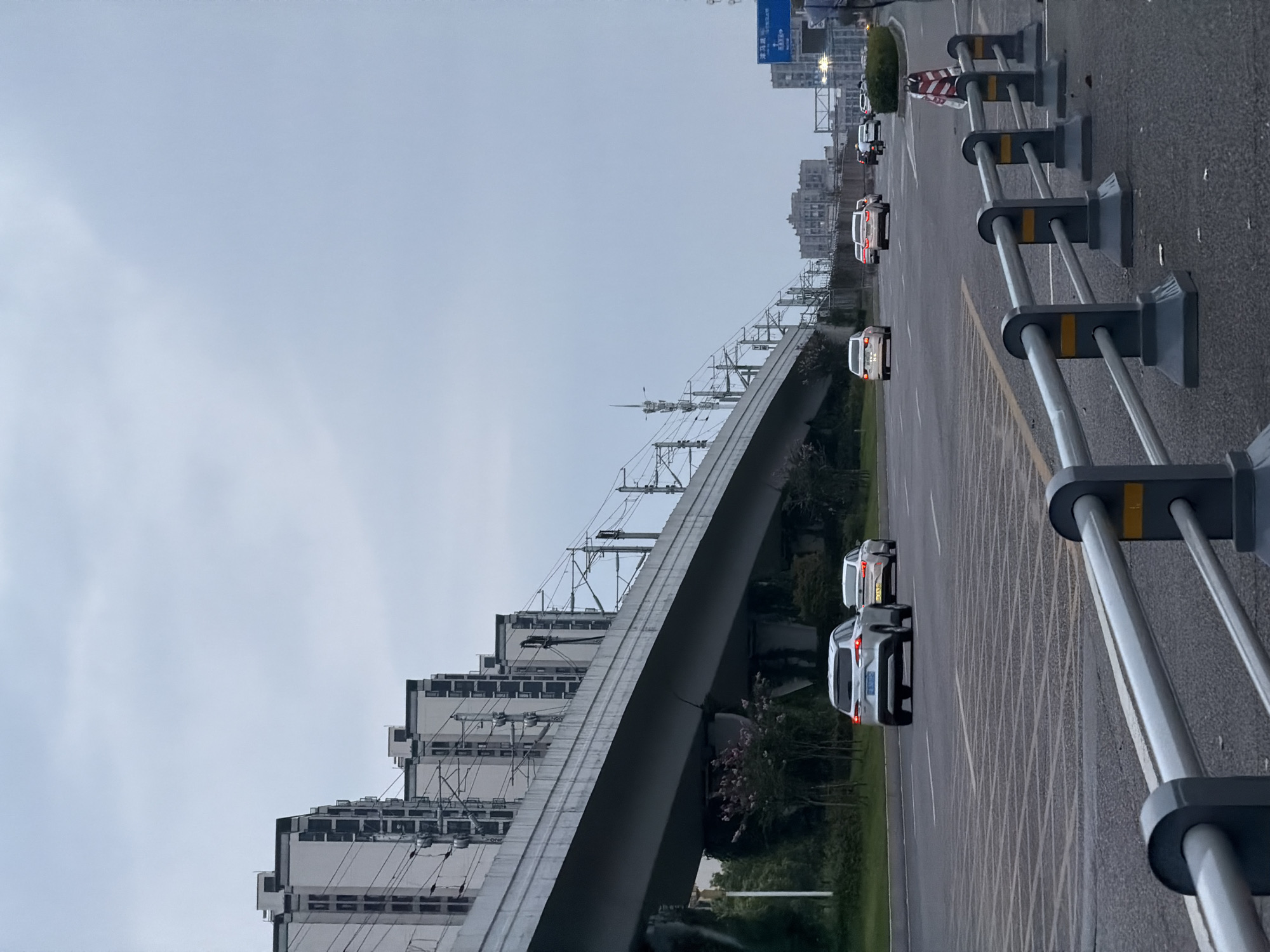 This is an eye-level shot of a multi-lane road in Jiangjin, China, with an elevated train track system running parallel to the road. The sky is overcast and light gray. The road is paved with asphalt and marked with white lane dividers and a rectangular grid pattern. Several cars are driving on the road, including a silver SUV in the foreground. On the left side of the road, there are several modern, multi-story buildings with repeating window patterns and black roofs, situated beneath the elevated train structure. The train tracks are supported by concrete pillars and feature a complex network of overhead electrical wires and poles. To the right of the road, there is a metal guardrail with gray posts and silver bars, interspersed with yellow reflective markers. Further down the road on the right, a blue traffic sign with Chinese characters is visible. Beyond the road and buildings, there are some trees and a slightly hazy view of other city buildings in the distance. A person in dark clothing is walking on the right edge of the road in the background.