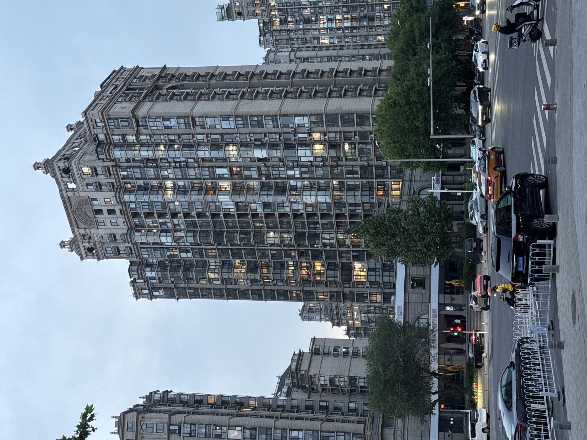 The image shows a street scene in Chaoyang District, China, featuring tall residential buildings and street-level activity. The buildings have a repeating window pattern and a light gray facade. Cars are parked and driving on the street. There are trees lining the sidewalk and traffic lights in the background. People are walking, and one person is riding a scooter.