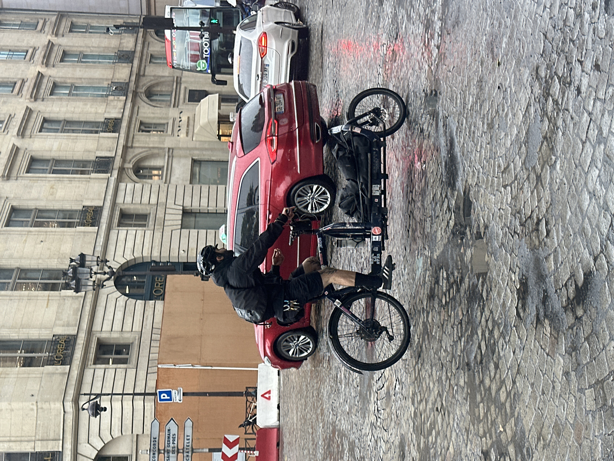 The image depicts a man on a bicycle in Paris, France. The man is wearing a black jacket, shorts, and a helmet, and he is riding a black bicycle with a trailer attached. The street is paved with cobblestones and is wet, suggesting it has recently rained.
Several vehicles are visible, including a red car, a white car, and a "TOOT BUS." The background buildings are ornate and typical of Parisian architecture, with signage indicating "L'OREAL." Street signs point to various destinations like "CONCORDE," "SAINT GERMAIN-DES-PRES," and "CHATELET." There is also a parking ("P") sign.