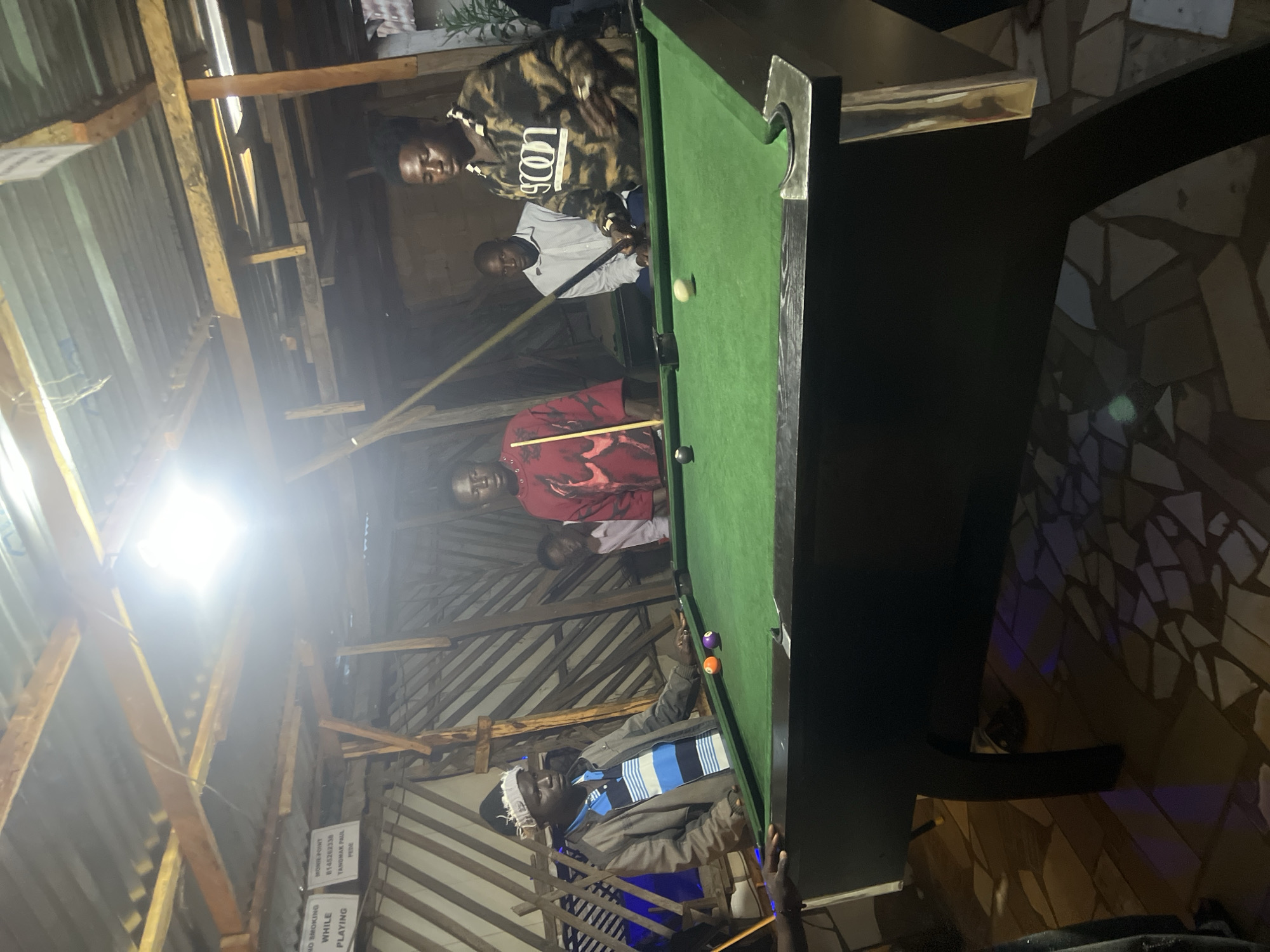 A group of men are gathered around a billiard table in what appears to be an informal establishment in Durumi, Nigeria. The setting is rustic, with exposed wooden beams and a corrugated metal roof, suggesting an outdoor or semi-enclosed space. The lighting is dim, with a bright overhead light illuminating the billiard table and some of the players.
The men are dressed in casual attire. One man in the foreground, wearing a grey jacket and a striped shirt, is positioned at the billiard table, holding a cue. Another man, wearing a red shirt with a graphic design, stands on the opposite side of the table, also holding a cue. To their right, two other men are visible, one in a light-colored shirt and the other in a camouflage-patterned jacket, both observing the game. Two billiard balls, one orange and one purple, are on the green felt of the table, as is a white cue ball.
The floor is paved with irregularly shaped tiles, some of which are illuminated by colored light, possibly from a nearby source not fully visible in the frame. There are signs visible on the wall in the background, one of which reads "NO SMOKING WHILE PLAYING." The overall impression is of a casual gathering for a game of billiards in a local setting.