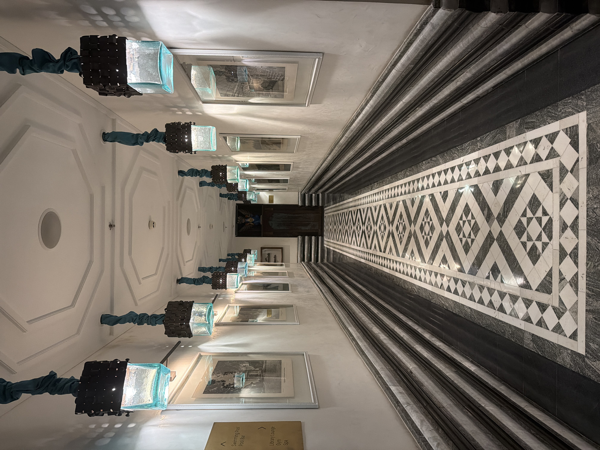 Here is a description of the image:
The image is a long, interior hallway shot with a vanishing point perspective. The walls are white, adorned with framed artwork and light fixtures featuring turquoise fabric and black lattice. The floor is a patterned black and white tile. A decorative, marble-like molding runs along the right side of the hallway. A sign is visible in the lower left. At the end of the hallway is a dark door with additional framed images. There are no people visible. The location is Unknown, Unknown.