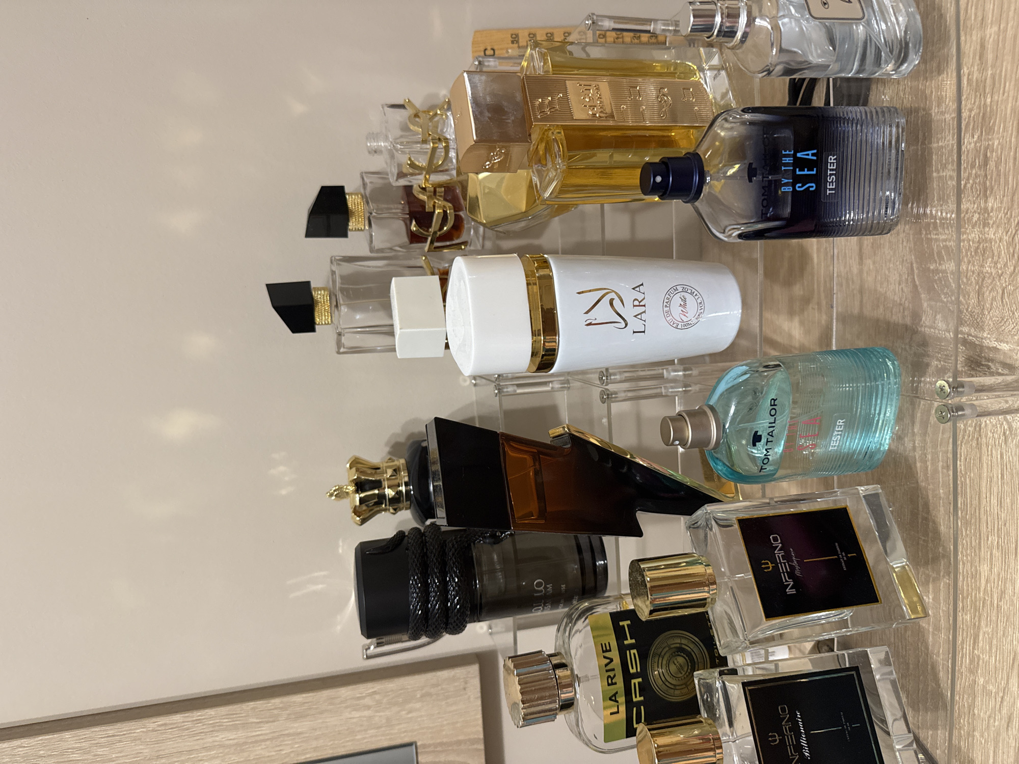 The image captures a collection of numerous perfume bottles meticulously arranged on clear, multi-tiered acrylic shelves. These shelves rest on a light-colored wooden surface, likely a dresser or shelf, against a plain, light-beige wall. No individuals are present in the scene.
The perfumes exhibit a wide array of bottle designs, sizes, and colors. Some bottles are transparent, revealing liquids in shades of golden-yellow or light blue, while others are opaque black or dark amber. Distinctive bottle features include a gold crown-shaped cap, a black bottle wrapped with a snake-like design, and several bottles adorned with elegant gold accents.
Several brand names and product details are visible. Text includes "LA RIVE CASH," "INFERNO Billionaire," and "INFERNO Masterpiece." Two "TOM TAILOR" bottles are present: one light blue and marked "TESTER," and another, a clear ribbed bottle with a dark blue label, titled "TOM TAILOR BY THE SEA TESTER." A white cylindrical bottle with a gold top prominently displays "LARA White EAU DE PARFUM 100ML." Another golden bottle features Arabic script, possibly "SANAM." A bottle bearing the "YSL" (Yves Saint Laurent) logo on its cap is also discernible. Faint numerical markings, such as "50," are visible behind some bottles, suggesting a ruler or measurement tool. The overall impression is an organized and personal display of fragrances.