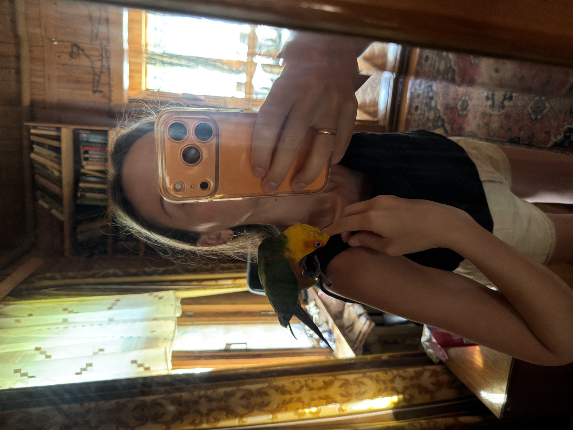 A young woman is taking a mirror selfie in a room with wooden walls and bookshelves. She is wearing a black tank top and beige shorts. A green and yellow parrot is perched on her shoulder and pecking at her finger. The room appears to be in a home in the city of Udelnaya, Russia, suggested by the rustic wooden interior and the presence of a bookshelf. The lighting suggests daytime.