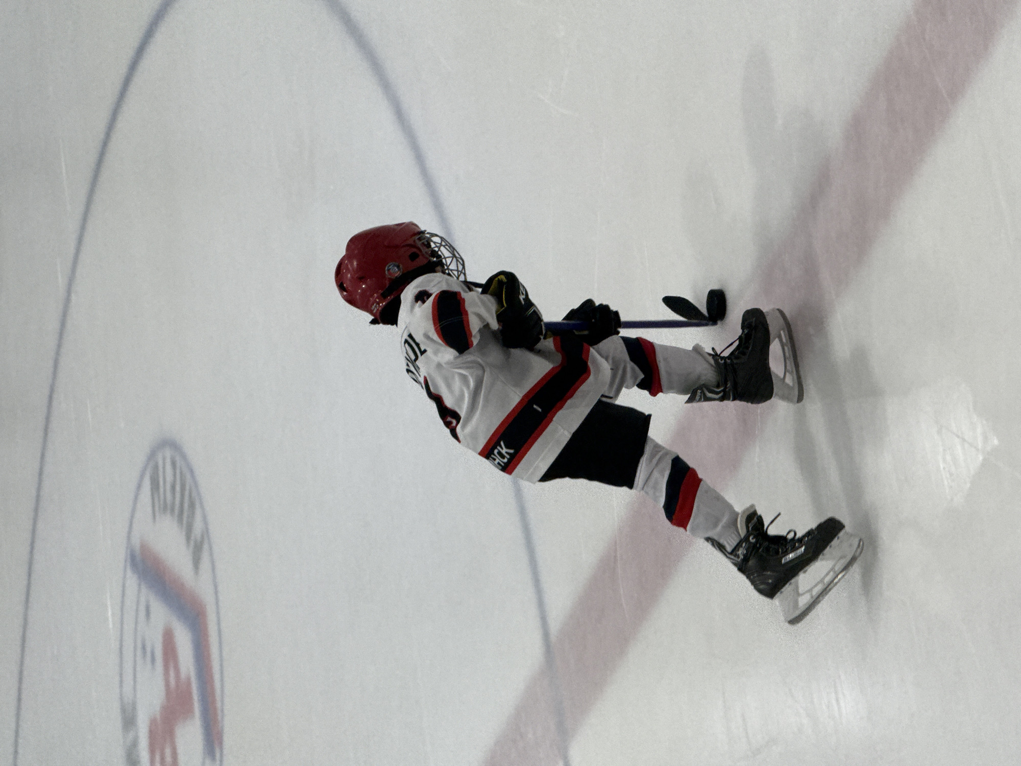 This media file shows a young person playing ice hockey on an ice rink in Votkinsk, Russia. The subject is wearing a white and red hockey jersey with black accents, black hockey pants, and a red helmet with a cage. They are holding a hockey stick and appear to be in motion. The ice rink has markings, including a partial logo of a circular emblem with red and blue elements. The lighting suggests it is daytime, likely indoors as there is no indication of natural weather conditions. The scene is focused on the action of the game, with the player captured in a dynamic pose. The visible text on the player's jersey reads "TCHOCK".