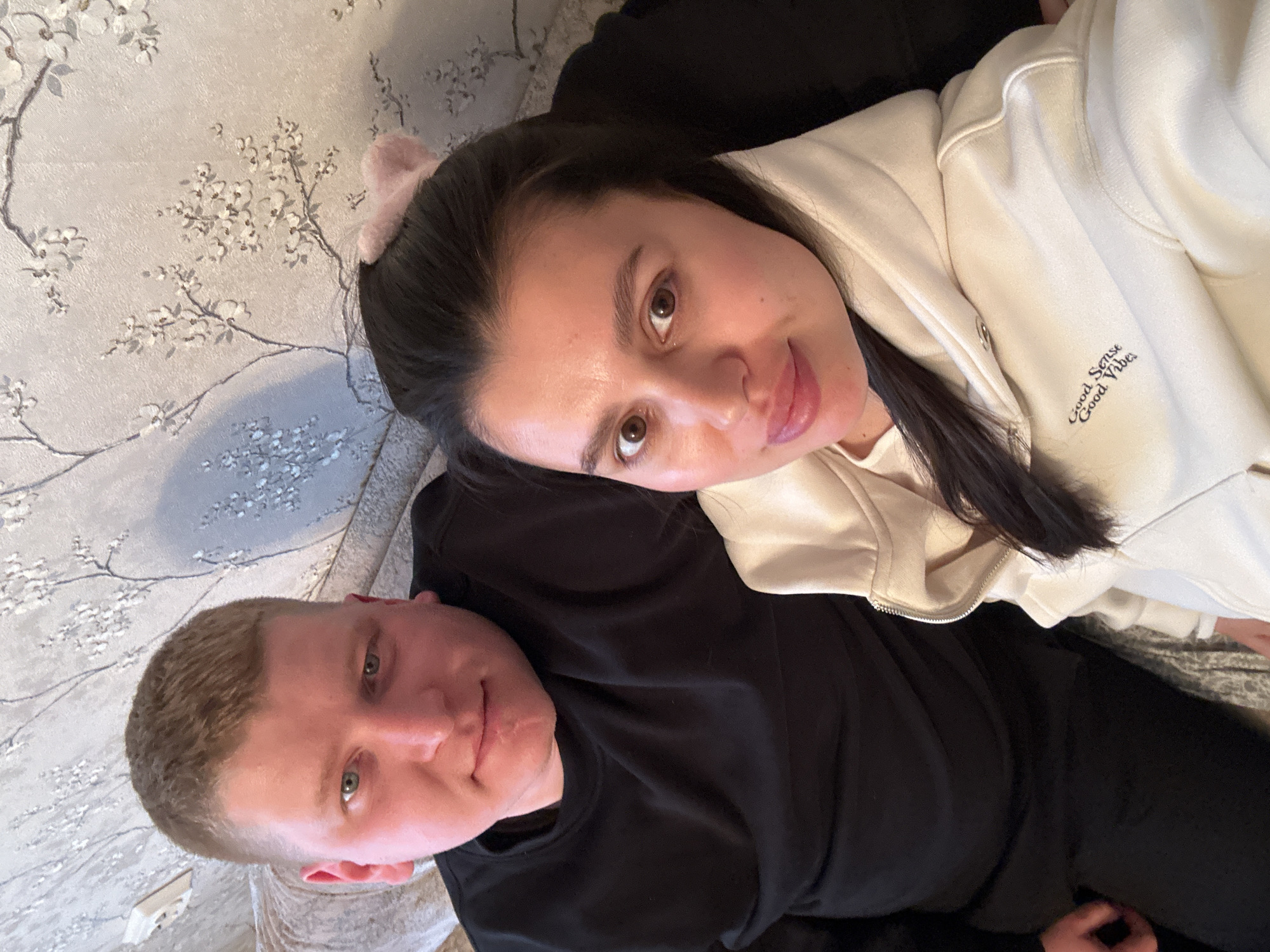 Two individuals are visible in an interior setting. A man with short, light-colored hair and a fair complexion is wearing a black sweatshirt. A woman with long, dark hair, light complexion and pink head band is wearing a cream-colored hooded sweatshirt with the words "Good Sense Good Vibes" embroidered on the sleeve. The woman is positioned above the man, and their heads are close together. The background includes a patterned wallpaper featuring floral designs and an electrical outlet. The location is specified as the city of Chaikovsky, Russia.
