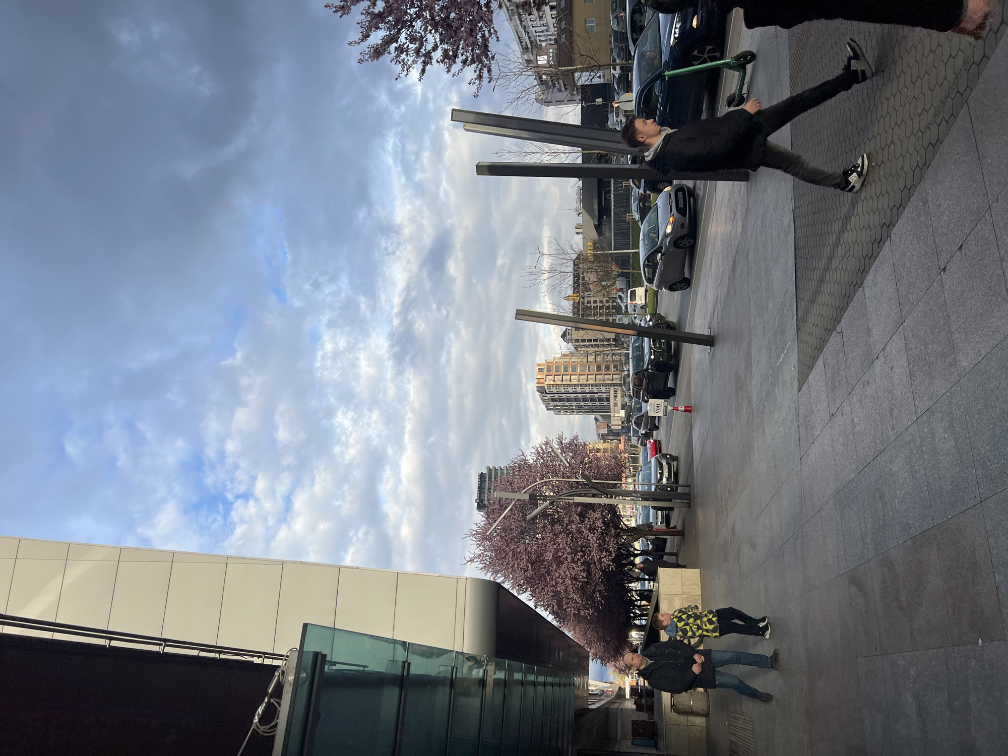 A medium shot captures a street scene in Sofia, Bulgaria, notable for its modern architecture and urban elements. The composition is viewed at an angle.
To the left, a contemporary building with glass and white paneling rises, its clean lines indicative of modern architectural design. Along the sidewalk, a row of blossoming plum trees adds a touch of nature to the urban landscape, their purple foliage contrasting with the gray tones of the surroundings.
Several individuals are present: one stands near metal poles, while another pair walks further down the street.
The right side of the image showcases a paved walkway, lined with parked cars. The sky above is filled with a mix of clouds and patches of blue.