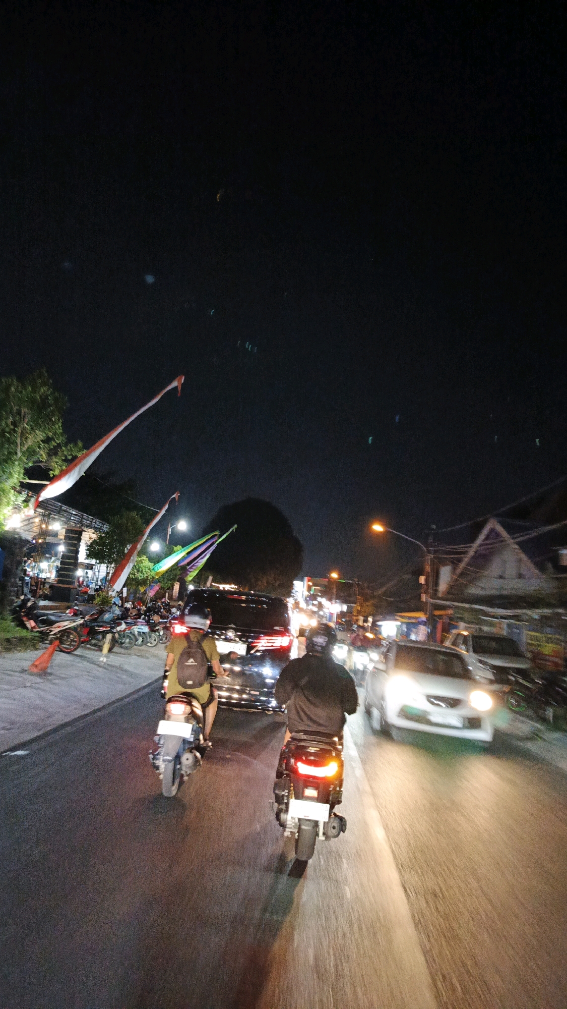 The image depicts a nighttime street scene in Surakarta, Indonesia, likely a busy thoroughfare with commercial establishments and heavy traffic. Two individuals on scooters are prominent in the foreground, traveling in the same direction. The scooter on the right is closer to the camera, with its rear brake light illuminated. The scooter on the left, slightly further ahead, has a rider wearing a backpack.
On the left side of the road, a row of shops or food stalls are visible, with lights illuminating the area and several parked motorcycles. Red and white flags, possibly national or celebratory, are prominently displayed, some stretched long across the street. Further down the street, colorful flags are also visible.
The road itself is paved and shows reflections from vehicle lights. Traffic includes cars and other motorcycles, with their headlights and taillights creating streaks of light due to the night conditions and possibly motion blur. A large, dark vehicle, possibly a van or SUV, is in the center lane, and a white car is in the right lane, its headlights bright. Overhead, streetlights and building lights illuminate the scene against a dark night sky, with some faint light flares visible. The overall atmosphere suggests a vibrant, active urban environment at night.
