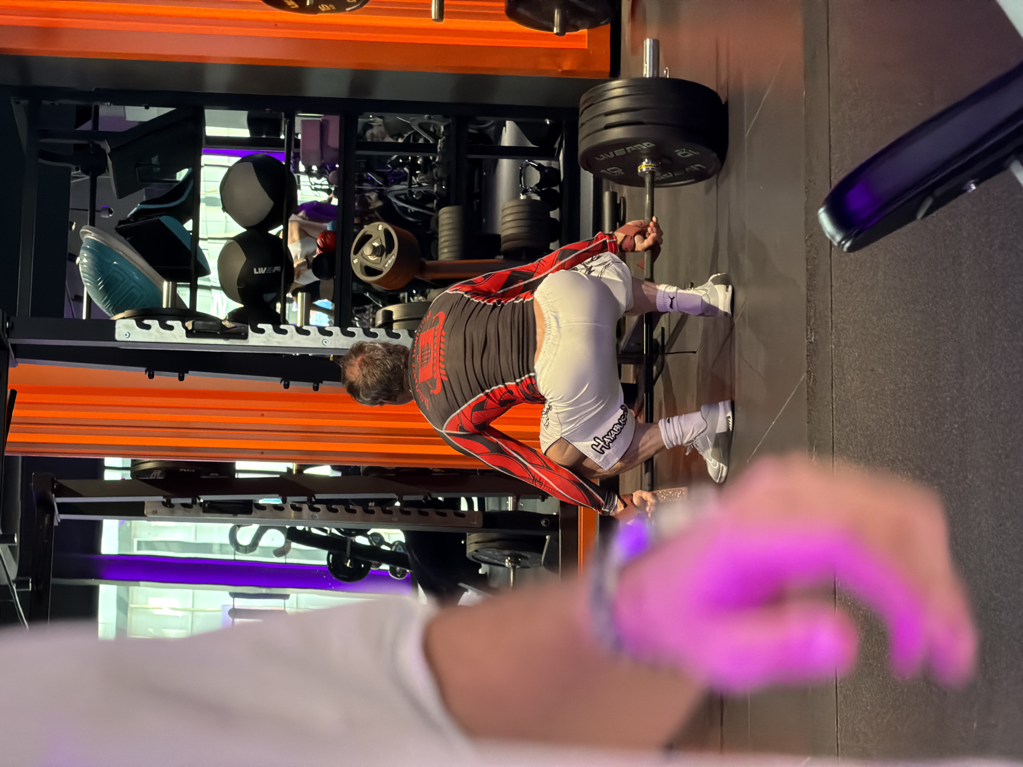 The image depicts a weightlifting scene, likely within a fitness center. An individual, a middle-aged man with graying hair, is positioned as if executing a barbell row exercise. He's wearing a black and red long-sleeved compression shirt, white shorts with "Hayabusa" written on them, white socks, and gray sneakers. He is holding a barbell loaded with black weight plates. The foreground is blurry and features a hand, likely holding a camera. The background includes a weight rack with various gym equipment, an orange stripe, and a metallic frame and gray walls. The scene doesn't provide sufficient detail to confirm it's in Saint Petersburg, Russia.