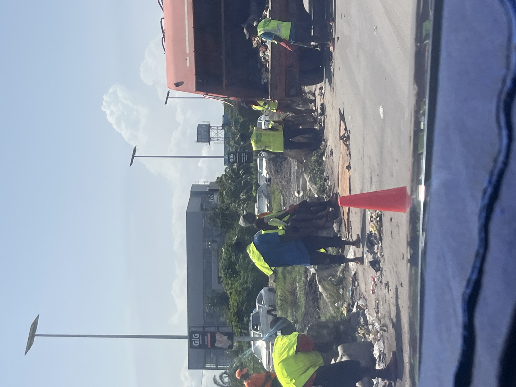 The image depicts a street scene in Lagos, Nigeria, characterized by garbage collection and street cleaning activities.
Several workers wearing high-visibility vests are present. Some are loading waste into the back of a truck. There is a lot of trash on the roadside.
A GIG billboard is visible. The sky is partly cloudy. Traffic cones are placed on the road to redirect traffic.