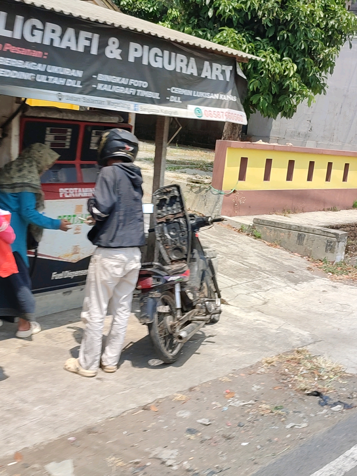 The image depicts a street scene in Salaman, Indonesia. In the foreground, a person wearing a black hoodie and light-colored pants stands next to a motorcycle. The motorcycle is parked on a concrete path. To the left of the motorcycle, a person wearing a blue shirt and a headscarf is interacting with another person who is partially visible. Behind them is a small shop with a black banner displaying text in white lettering, which appears to be a business sign. The banner advertises "LIGRAFI & PIGURA ART" and lists services such as "BERBAGAI UKURAN" (various sizes), "BEDDING ULTAH DLL" (birthday bedding, etc.), "- BINGKAI FOTO" (photo frames), and "- KALIGRAFI TIMBUL DLL" (embossed calligraphy, etc.). Below the banner, there is a sign with the phone number "08587XXXXXX". The shop has a red and white exterior. In the background, a yellow wall with brown vertical accents is visible. The environment appears to be an outdoor urban setting with trees and foliage in the upper portion of the image. The ground is paved with concrete and scattered with debris. The overall impression is that of a street-side business operating in a somewhat modest urban environment.