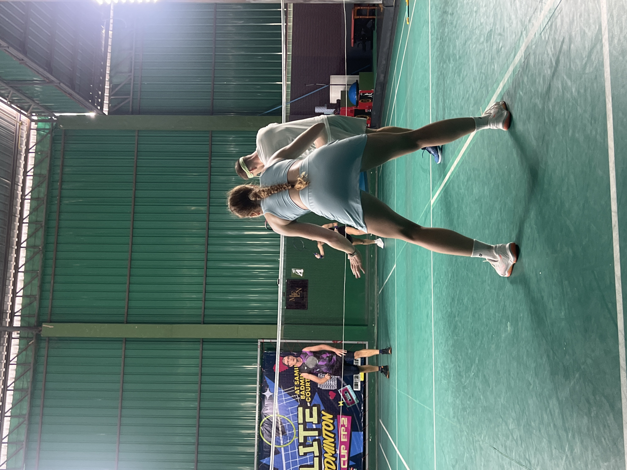 A group of four individuals are playing badminton indoors on a green court. In the foreground, a woman with a braided ponytail is facing away from the camera, wearing a light blue athletic outfit and white socks with orange-soled tennis shoes. To her right and slightly behind her, a man with a green headband is wearing a light blue t-shirt and shorts. Further back, a man in a dark purple tank top and black shorts, wearing a red baseball cap, stands near the net. Another person, a woman, is visible further in the background. The court is surrounded by green walls and a metal roof structure. A banner with text, including "LITE" and "BADMINTON CUP EP.2," is visible on the left side of the court. The lighting suggests it is daytime, possibly with artificial lights supplementing natural light.