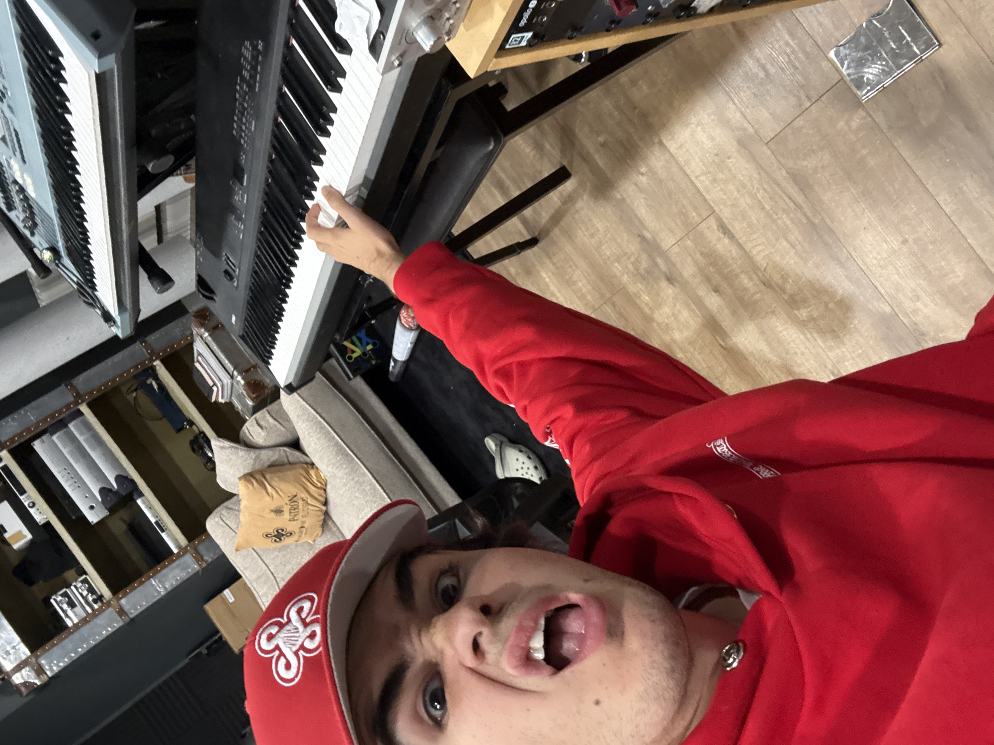 A young person wearing a red hoodie and a red baseball cap with a white logo is taking a selfie in a room with musical instruments and shelves. The person has a surprised or exaggerated expression, with eyes wide open and mouth agape. Their right hand is extended, with fingers touching the keys of a black keyboard. Behind them, there is a beige sofa with a small, orange pillow that has the Patron logo on it. Two keyboards are stacked on a stand, one blue and one black, with the person's hand on the lower black keyboard. Shelves filled with audio equipment and boxes are visible in the background. The floor is made of light brown wooden planks. A gray Croc shoe is on the floor near the sofa. There is also a piece of crinkled foil on the floor. The scene appears to be an indoor music studio or practice space.